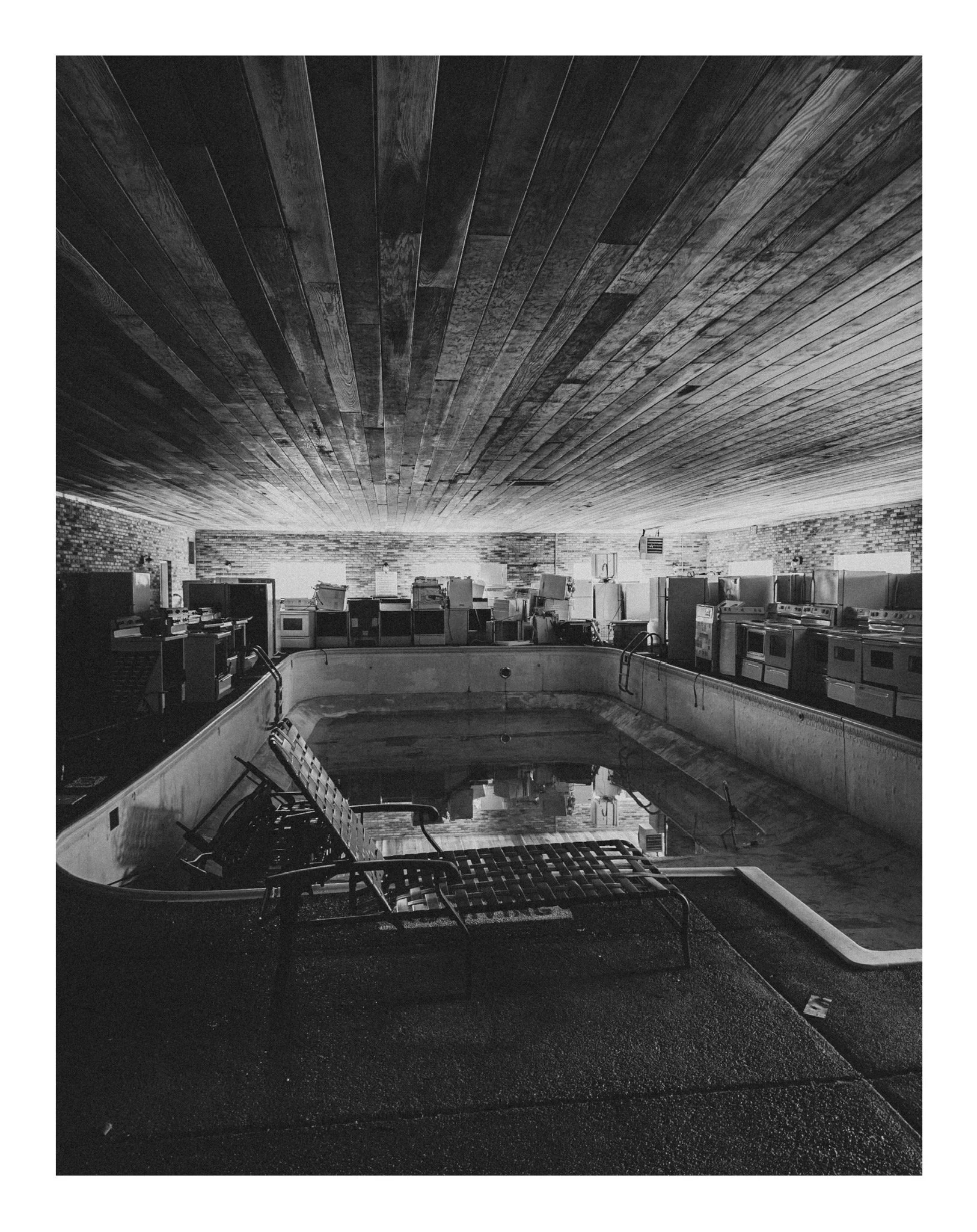 An indoor swimming pool area with empty lounge chairs, a brick wall, pool ladder, and a patterned ceiling.
