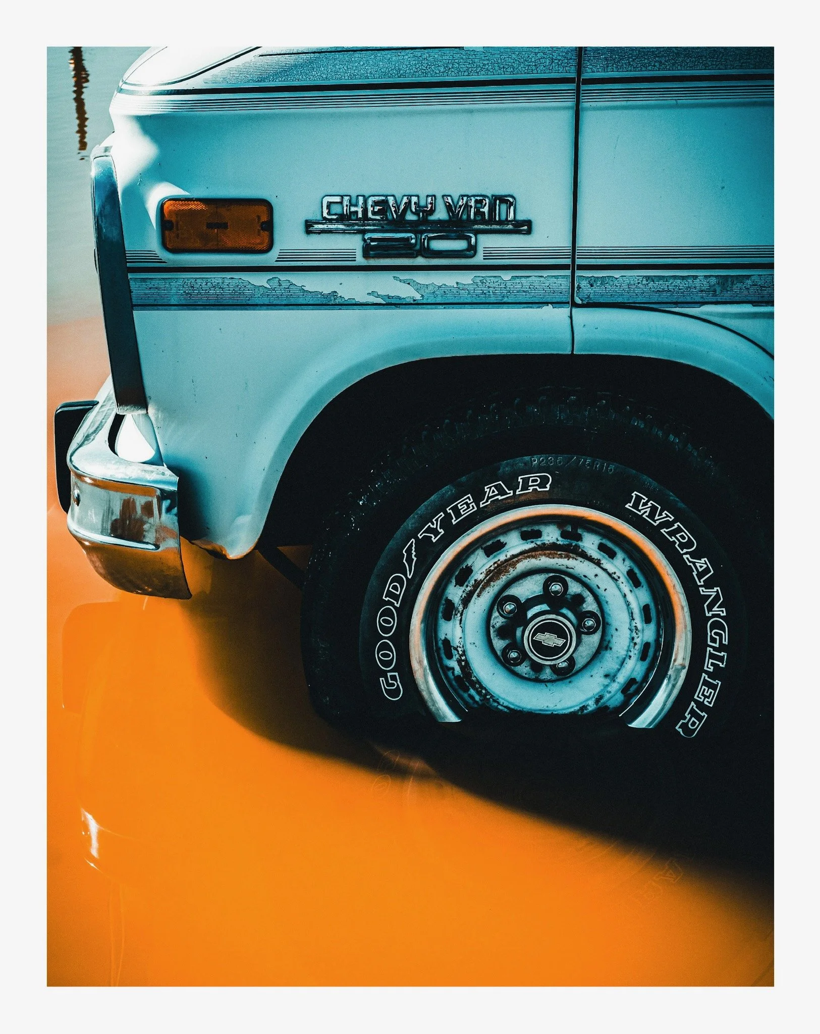 Close-up of the front side of a vintage Chevrolet truck, showing the Chevrolet emblem and part of the wheel with Goodyear Wrangler tire, against an orange background.