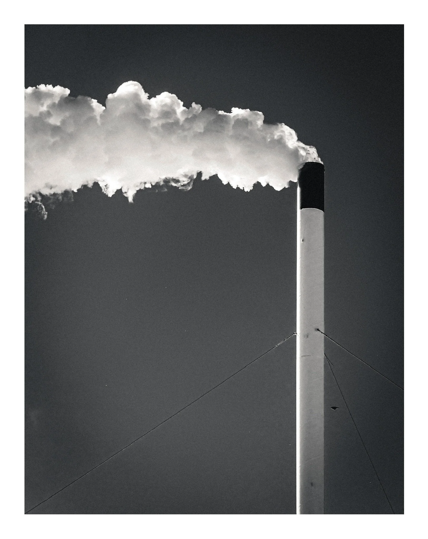 A tall smokestack or chimney emitting a large cloud of smoke into the sky, with supporting cables attached to the structure.