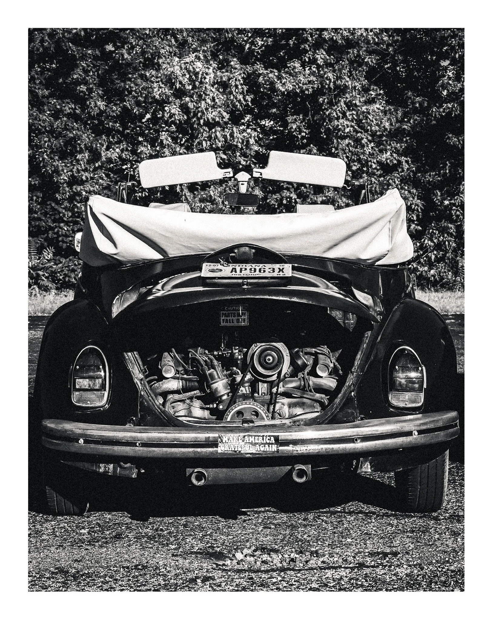 Black and white image of a car with an open hood, showing the engine. The car has a license plate reading 'AD963X' from Indiana, and a bumper sticker that says 'Make America Grateful Again.' The car's windshield has two flip-up sun visors, and a clot