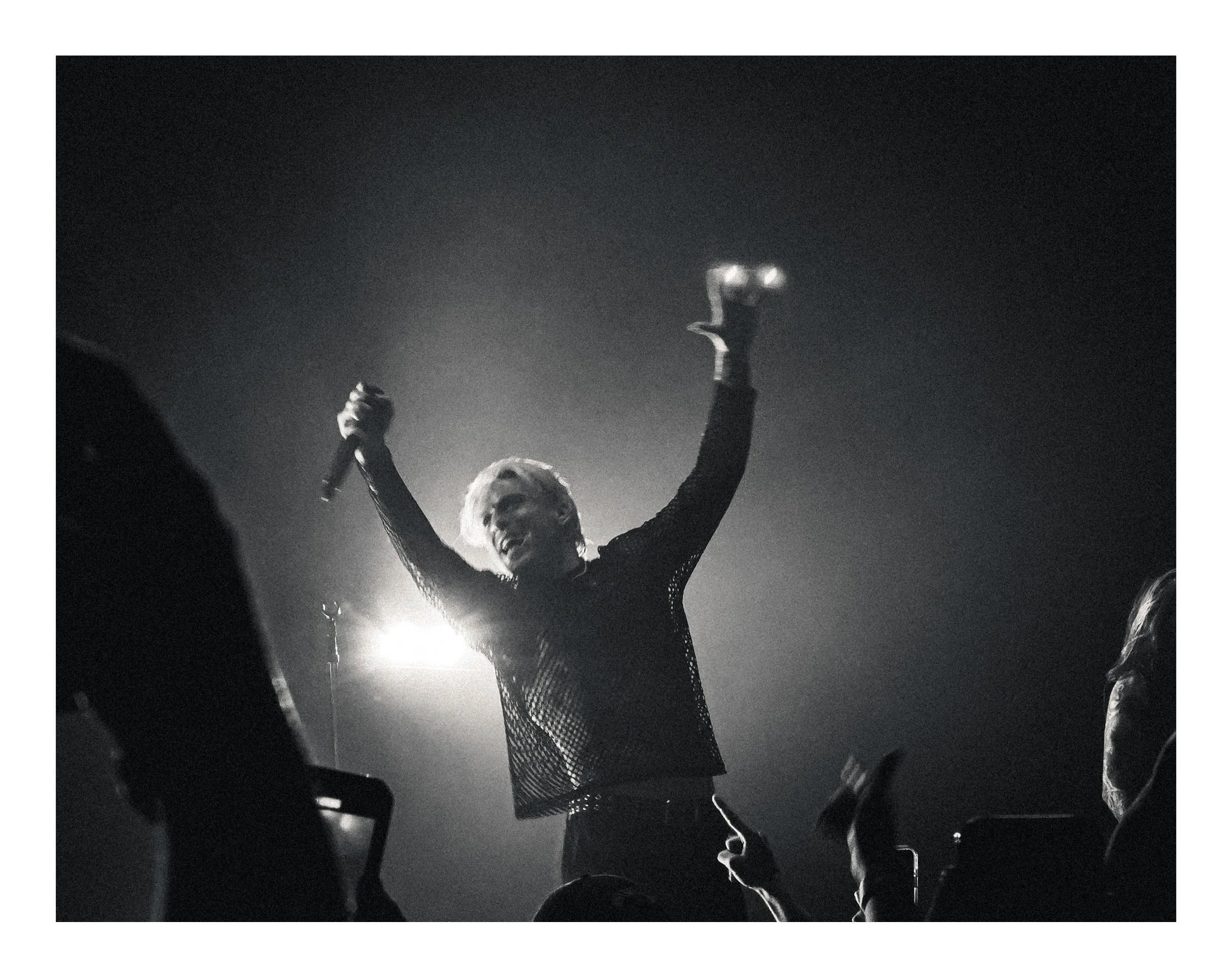 A performer on stage with arms raised, holding a microphone in one hand, illuminated by stage lights, wearing a fishnet top, and surrounded by an audience in black and white.