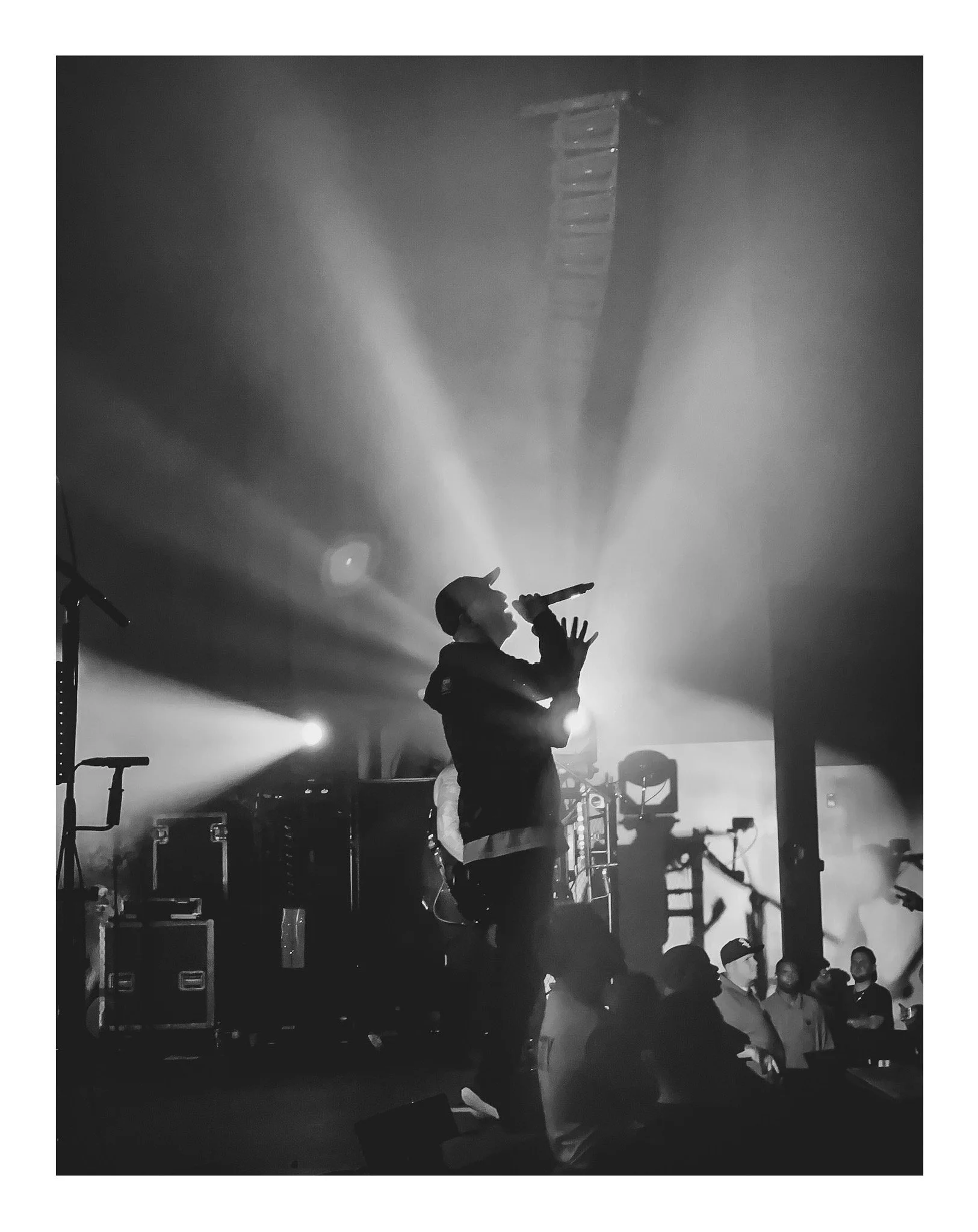 A singer performing on stage with a microphone, surrounded by stage equipment, with a group of people watching in the foreground.