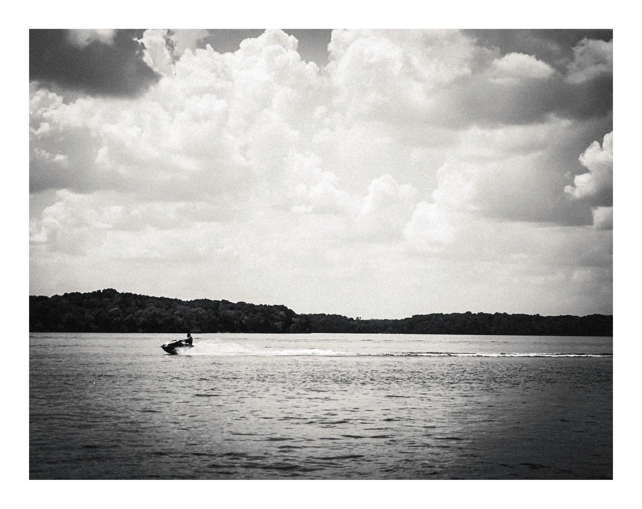 Black and white photo of a person riding a jet ski on a body of water, with a distant tree-lined shoreline and cloudy sky.