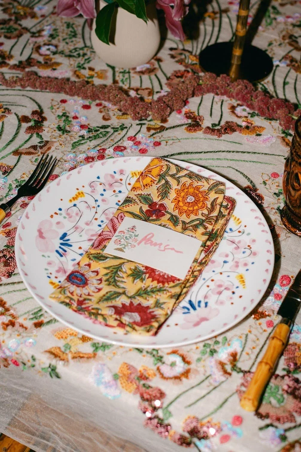 A table setting with a floral napkin, a decorative plate, a place card with the name 'Paige', and floral embroidered tablecloth with a vase and candlestick in the background.