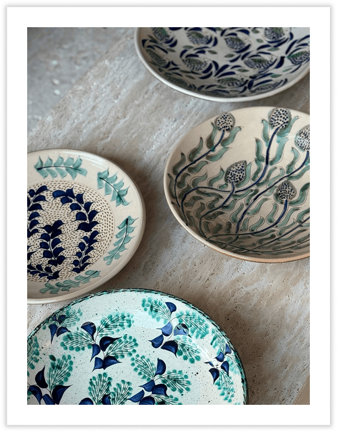 Four decorative ceramic plates with floral and botanical designs in blue, green, and black, placed on a light-colored surface.