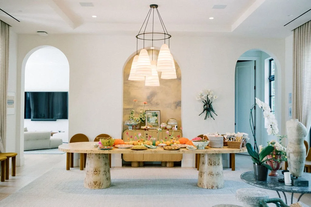 A dining room with a large wooden table set with colorful dishes, surrounded by chairs, with decorative flowers on the walls and a sideboard against the wall, and a modern chandelier hanging above the table.
