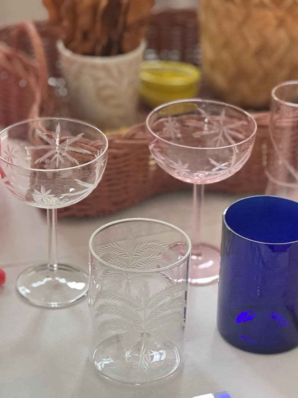 Set of glassware, including pink coupe glasses, a clear tumbler, and a blue cup, all with etched leaf and fern designs, on a table with a wicker basket and other items in the background.