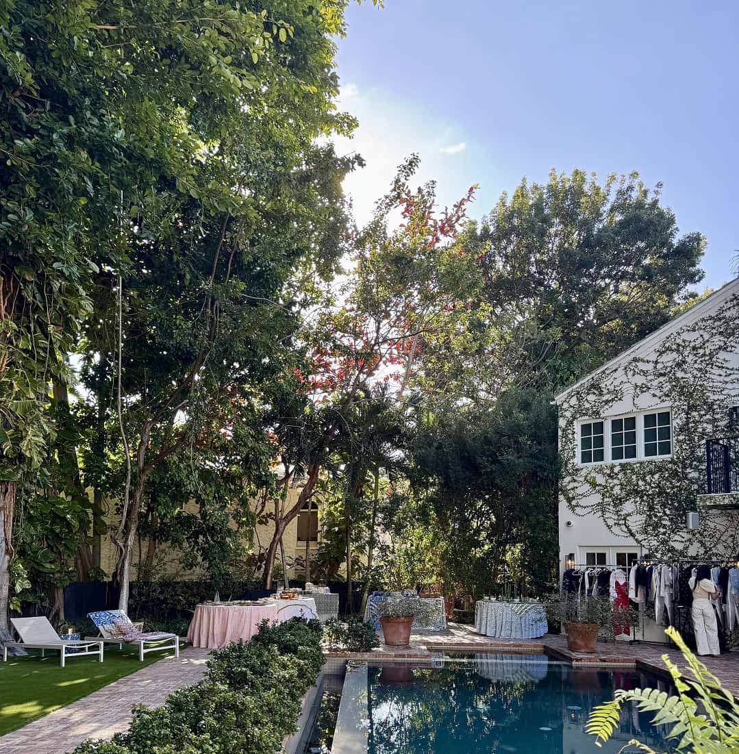 Backyard scene with a swimming pool, surrounded by lush green trees, and a white house with ivy climbing its walls. There are lounge chairs, tables with tablecloths, and outdoor clothing racks.