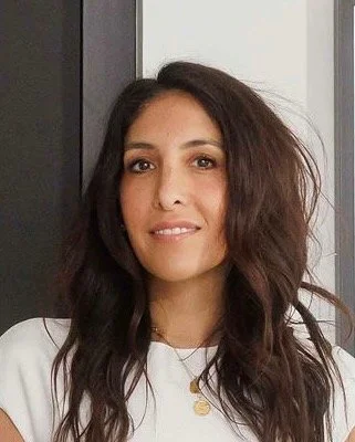 A woman with long, wavy dark hair, wearing a white top and gold necklace, standing indoors.