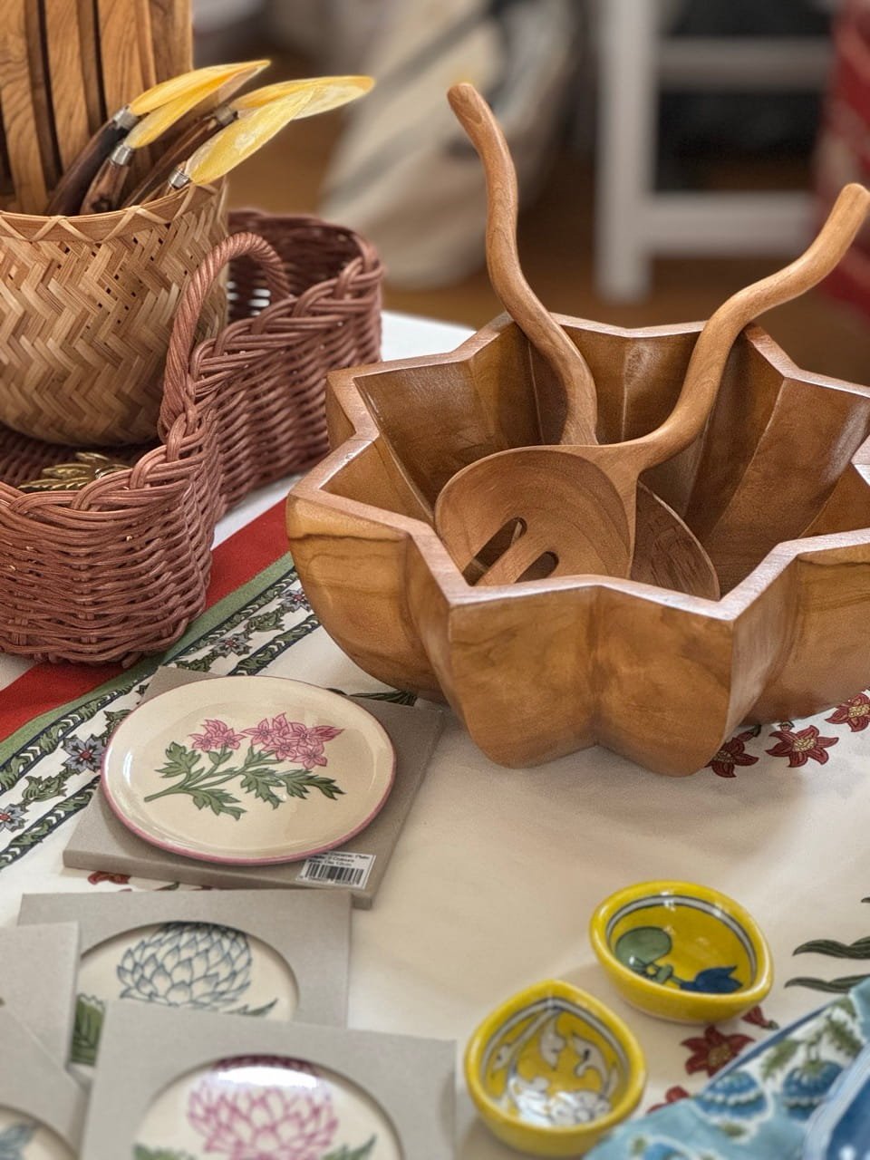 Display of handcrafted wooden bowls, ceramic plates with floral and nature designs, woven baskets, and small ceramic dishes with painted fruit on a table.