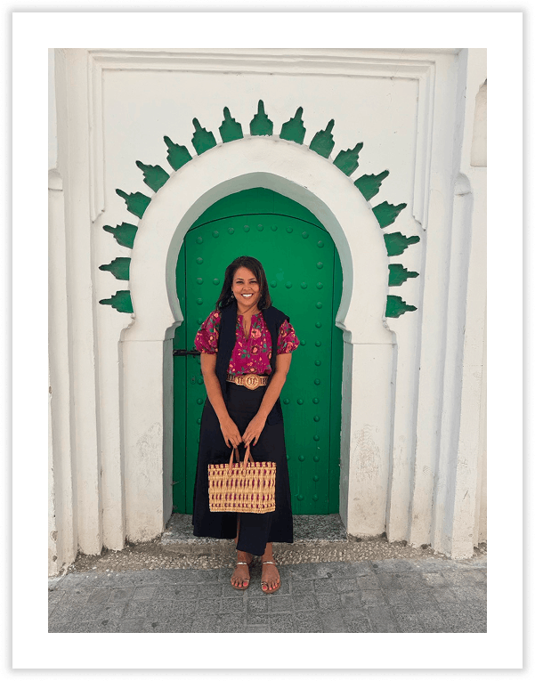 Woman standing in front of a white wall with a green arched door and green decorative elements above the door, smiling and holding a woven basket.