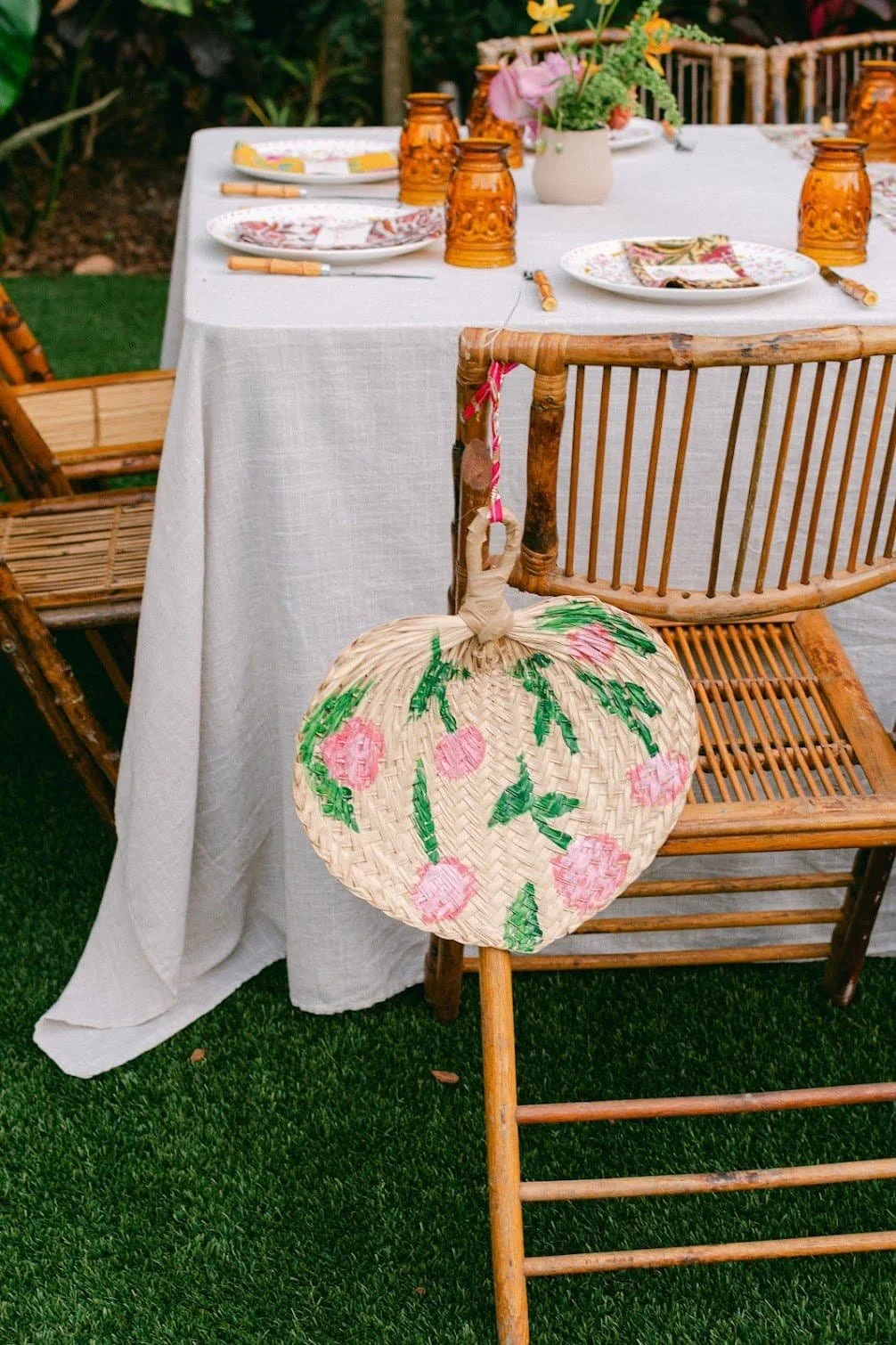 An outdoor dining table set with a white tablecloth, decorative amber glass lanterns, floral plates, and cutlery. A wicker chair and a hammock with a floral pattern hang from the back of the chair, with green grass underneath and lush greenery in the background.