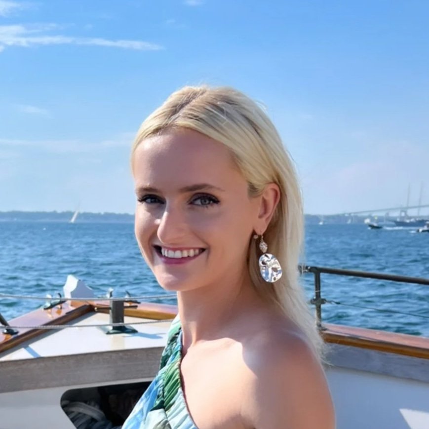 A smiling woman with blonde hair in a strapless dress standing on a boat with a blue ocean and a distant bridge in the background.