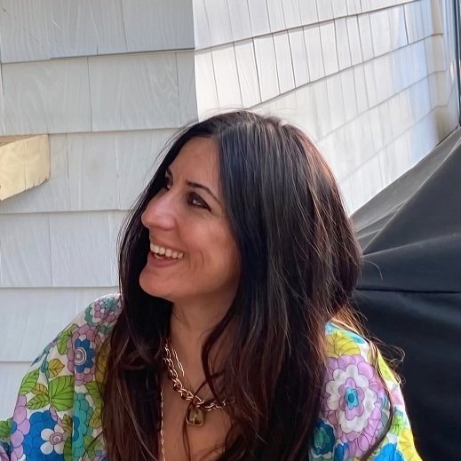 A woman with long dark hair smiling and looking to the side, wearing a colorful floral blouse and a gold chain necklace, standing outside near a house with siding.