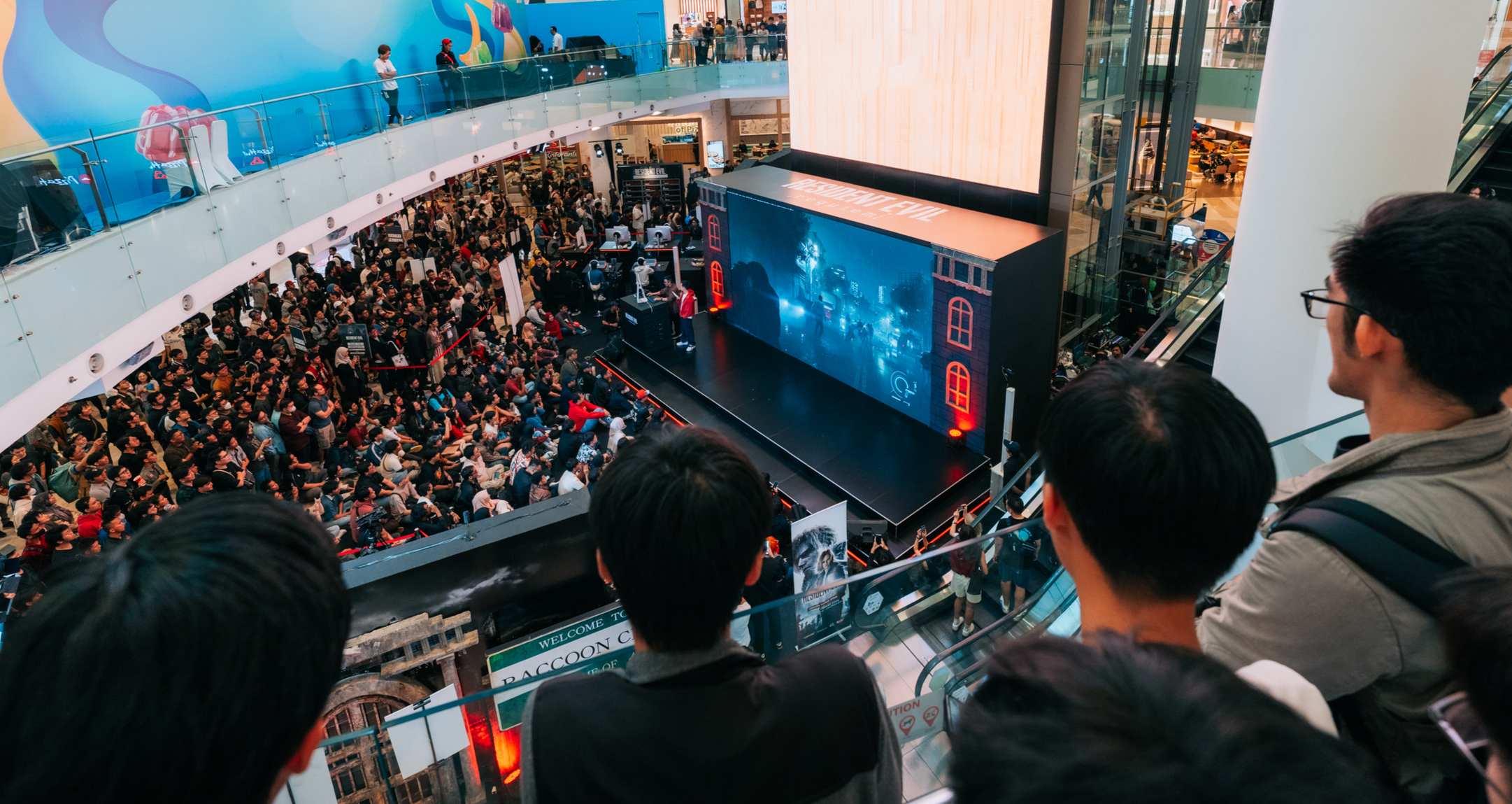 People gathered in a shopping mall's atrium watching a large screen display, with multiple levels visible and a crowd of onlookers.