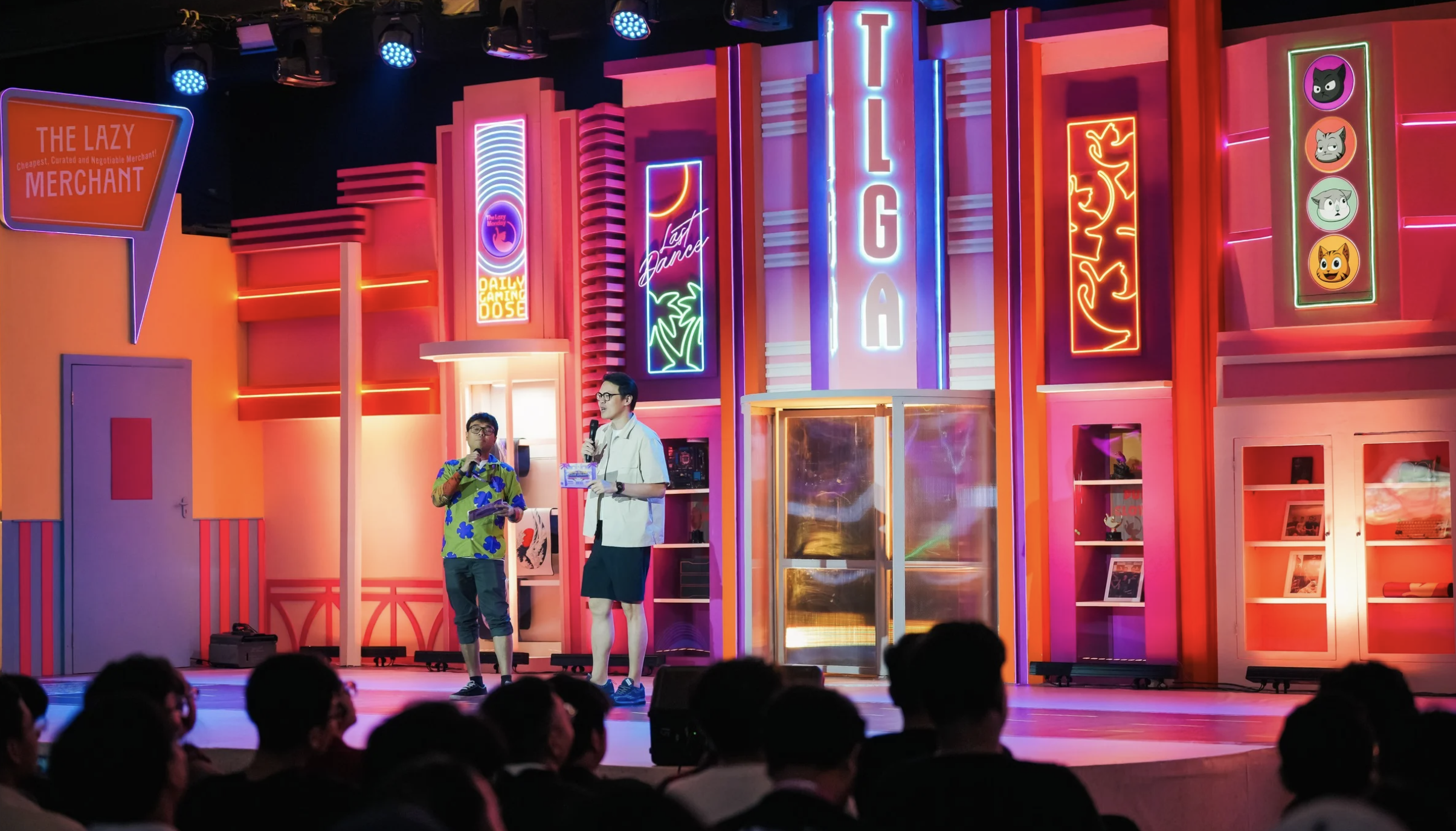 Two men speaking on stage in front of colorful, neon-lit signs and a vibrant background, with an audience watching.