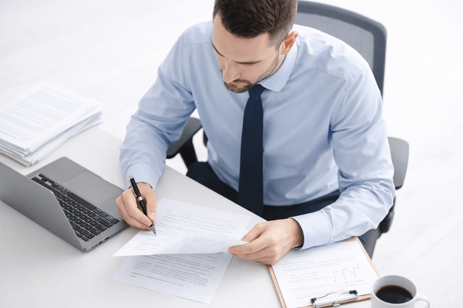 A man in a light blue shirt and dark tie is sitting at a desk, reviewing documents with a pen in his hand. There are stacks of papers, a laptop, a clipboard with graphs, and a cup of coffee on the desk.