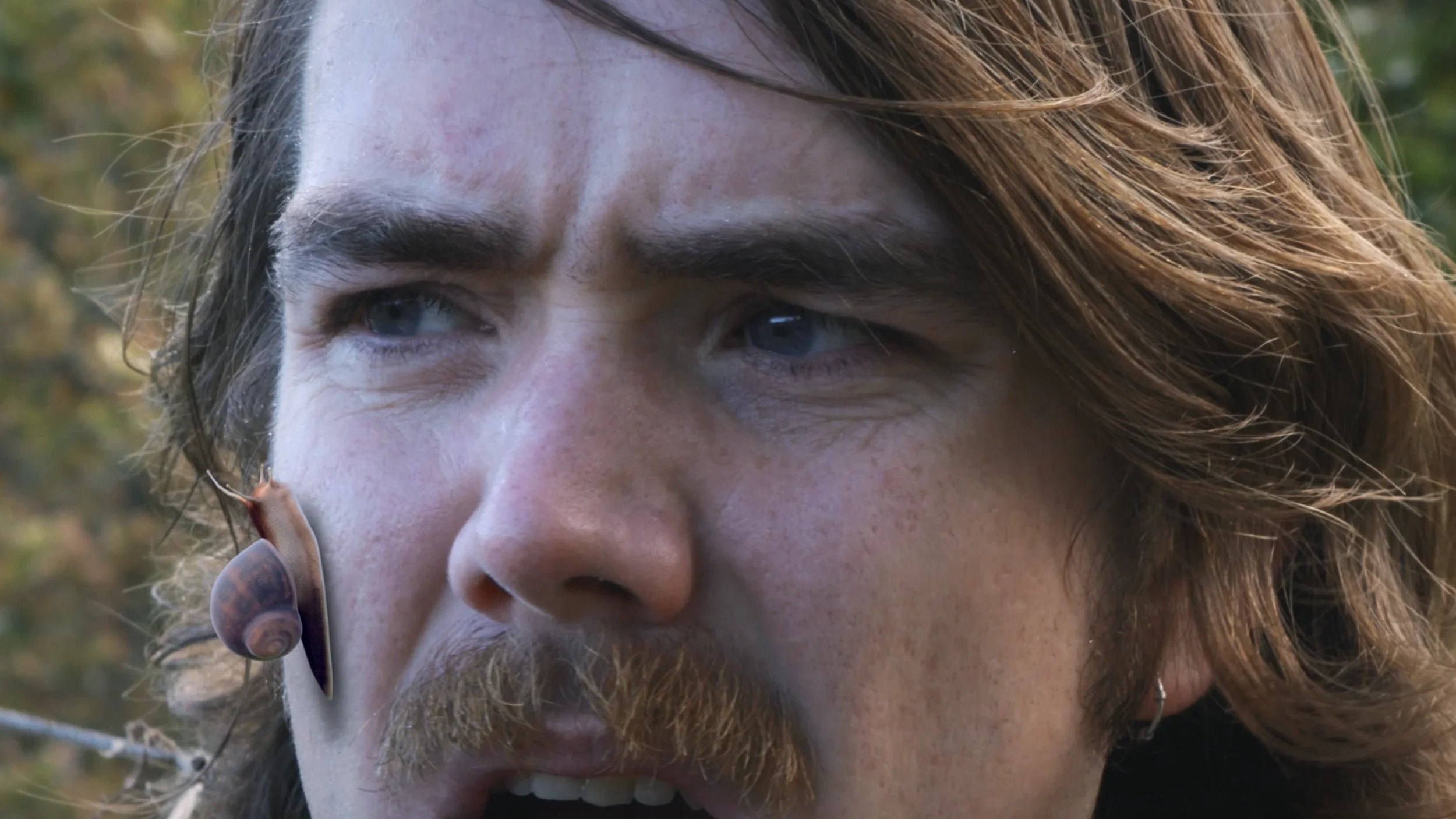 Close-up of a man with brown hair and mustache, wearing a snail-shaped earring on his left ear, outdoors with blurred greenery in the background.