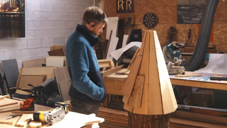 A man is working on a woodworking project in a workshop, carving a large wooden cone onto a tree trunk.