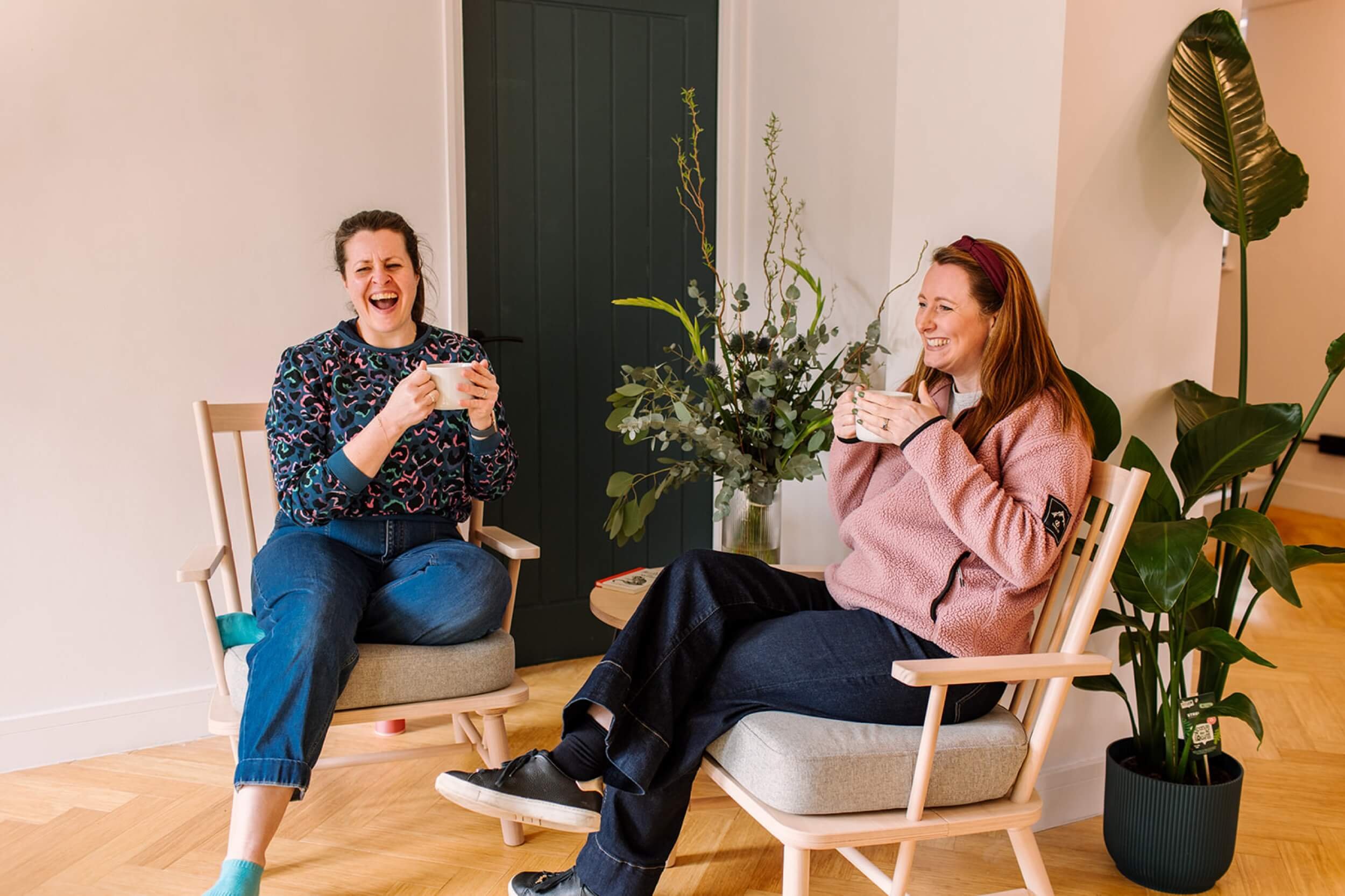 Two women sitting on wooden chairs, laughing and holding mugs, with a large green plant and floral arrangement between them in a cozy room.