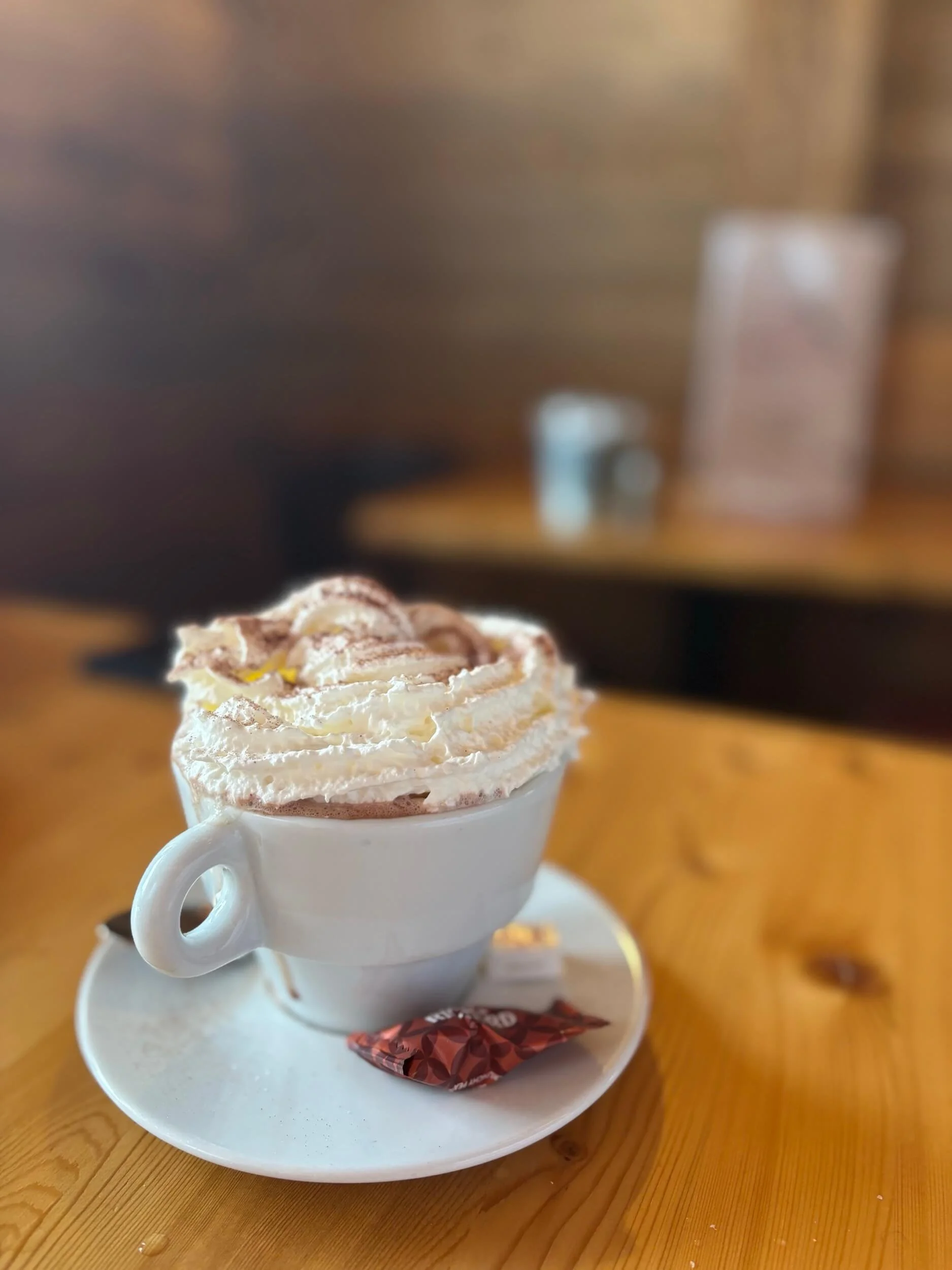 A cup of whipped cream-topped coffee or hot chocolate with cocoa powder, served on a small plate with a wrapped chocolate piece, on a wooden table.