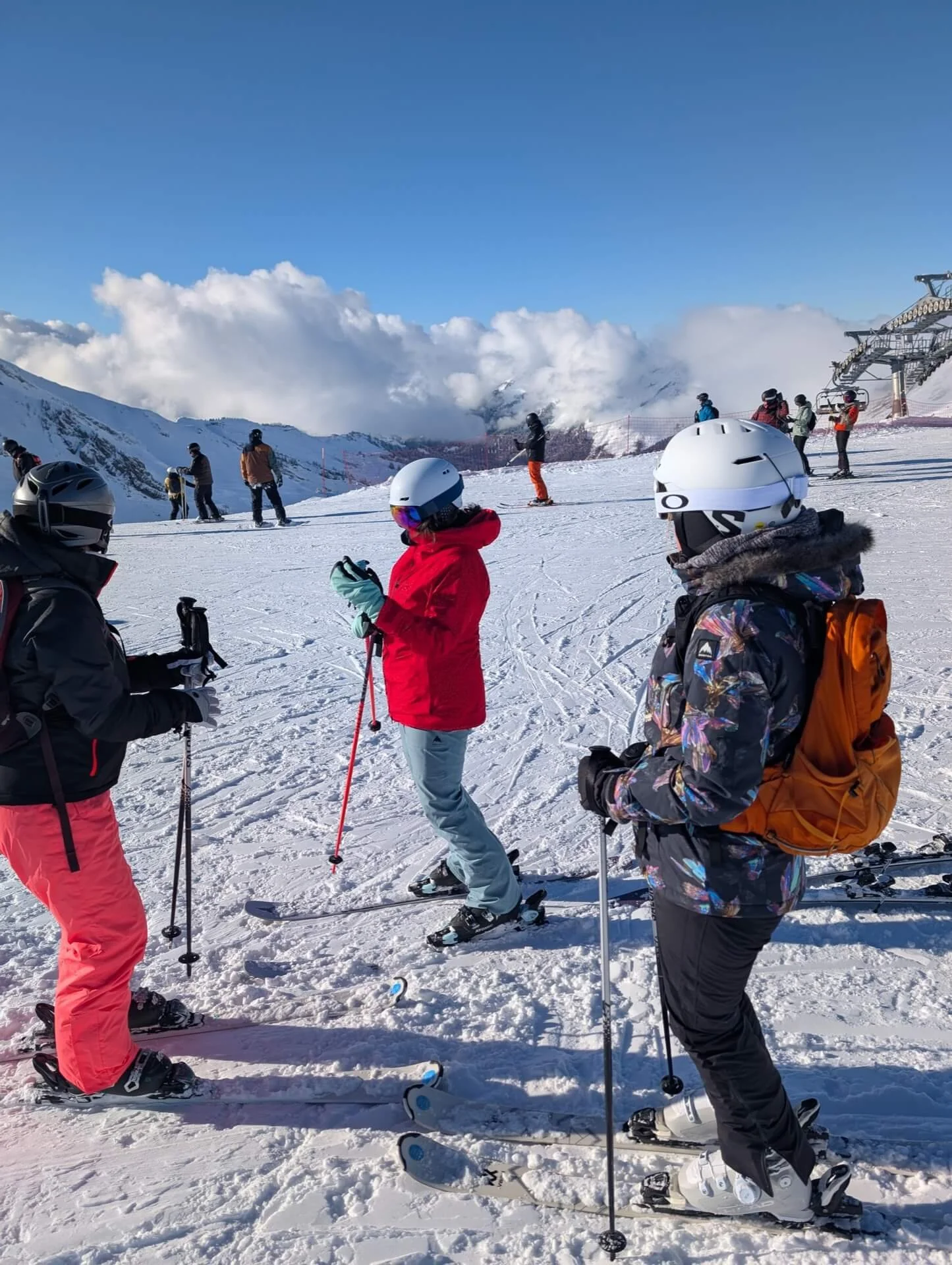 Group of skiers and snowboarders on a snowy mountain under a blue sky with clouds, some standing, some skiing or snowboarding, and ski lift in the background.