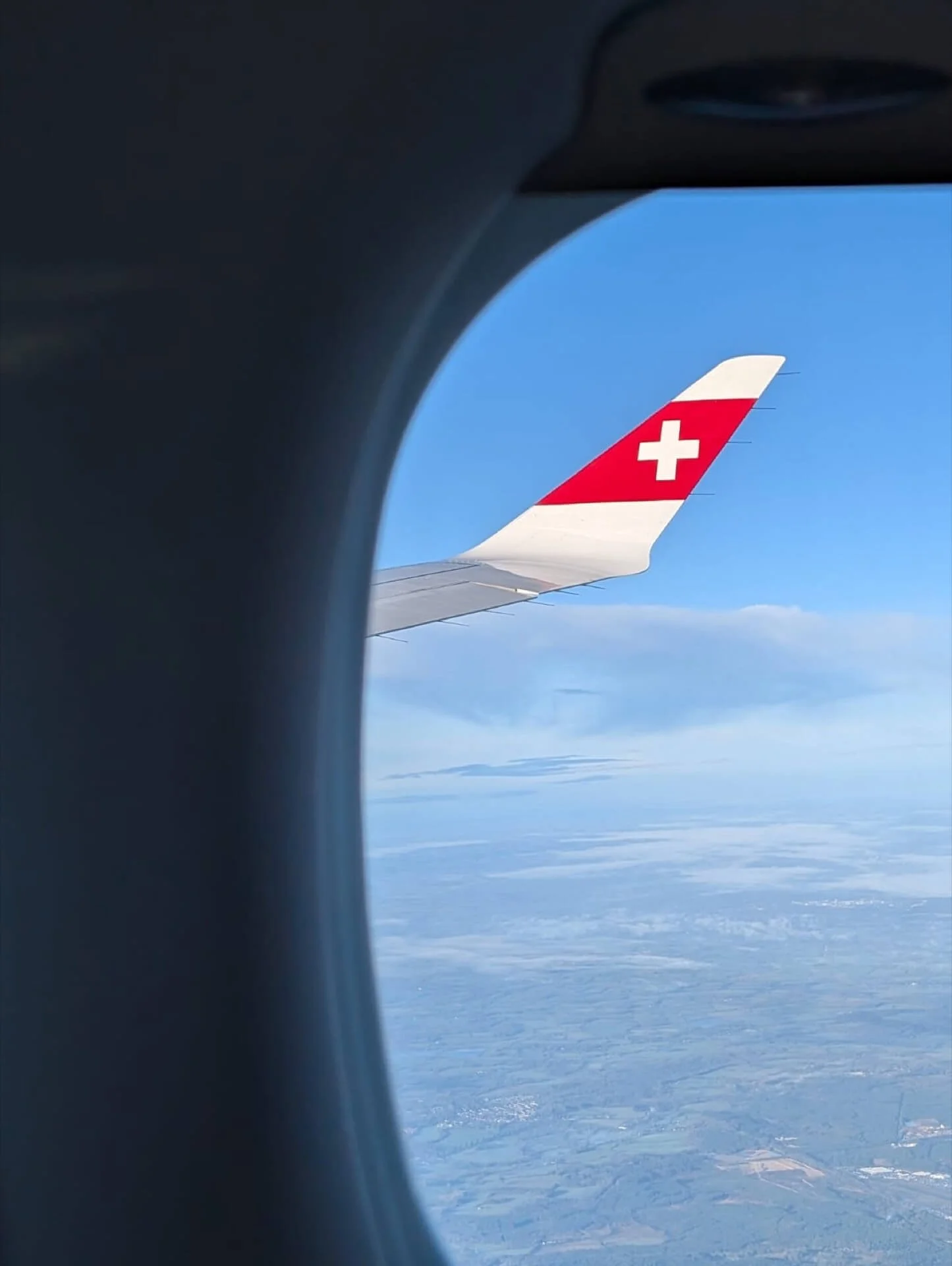 Photo taken from an airplane window showing the wing with a red and white Swiss Airlines logo against a blue sky and clouds below.