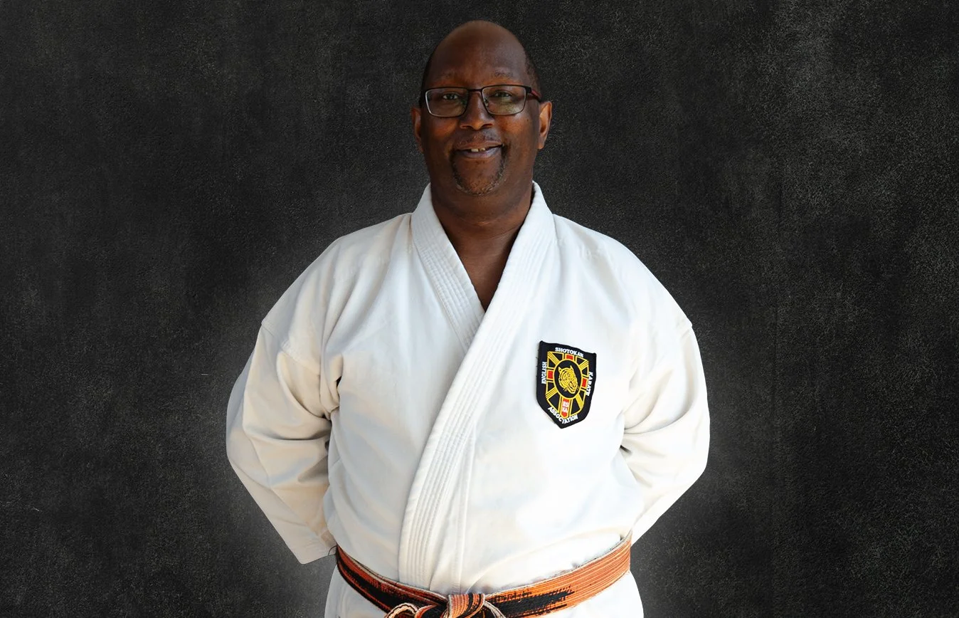 Sensei Wayne James, wearing a white martial arts gi with a black belt, standing against a black background.