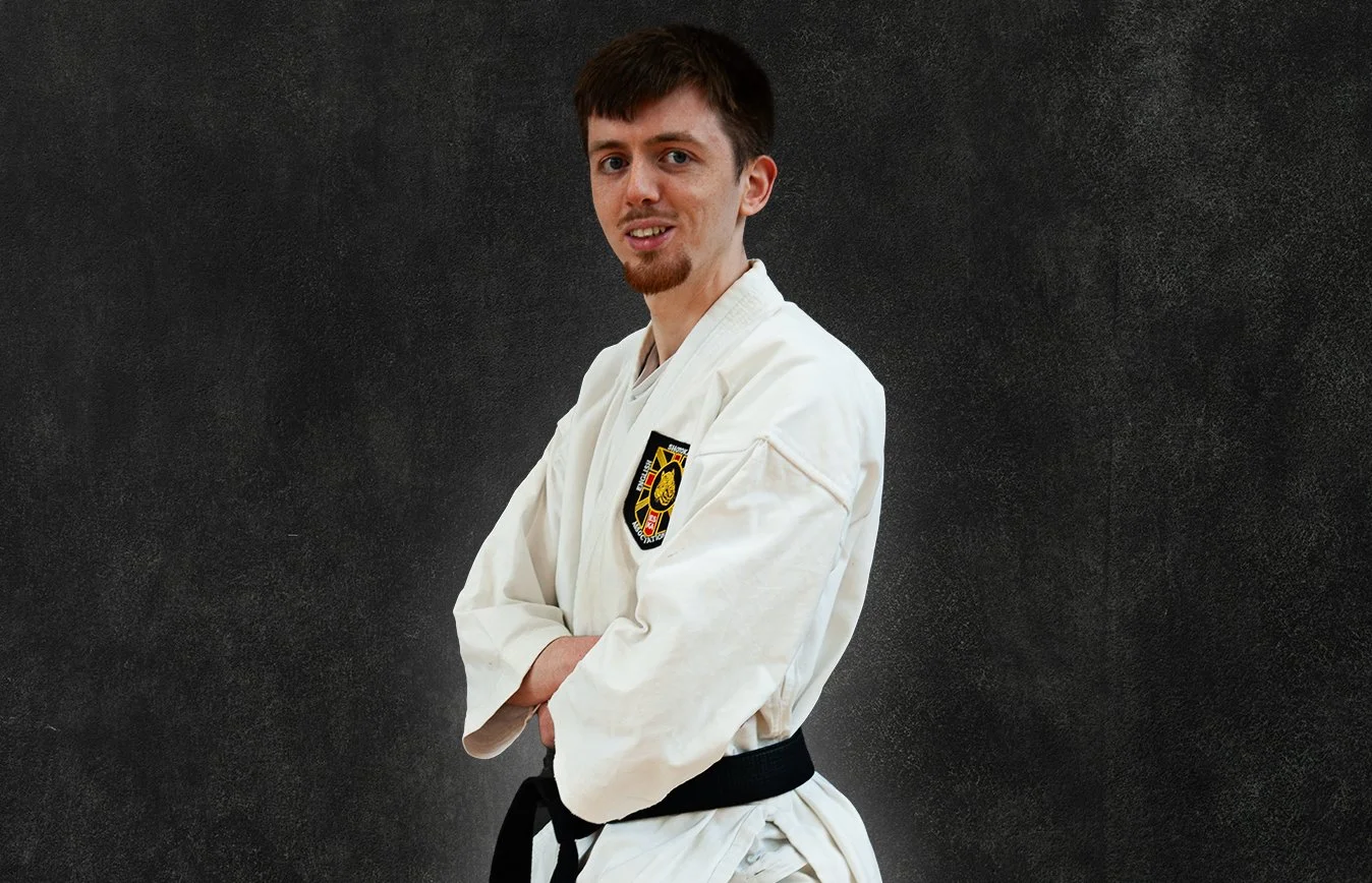 James Copland, wearing a white martial arts gi with a black belt, standing against a black background