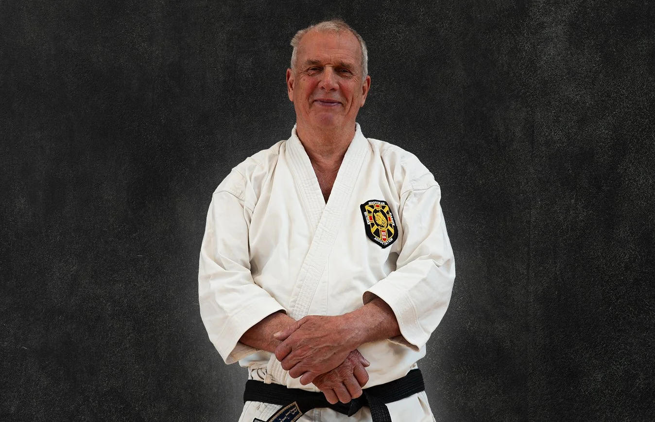 Sensei Angus Pearson, wearing a white martial arts gi with a black belt, standing against a black background