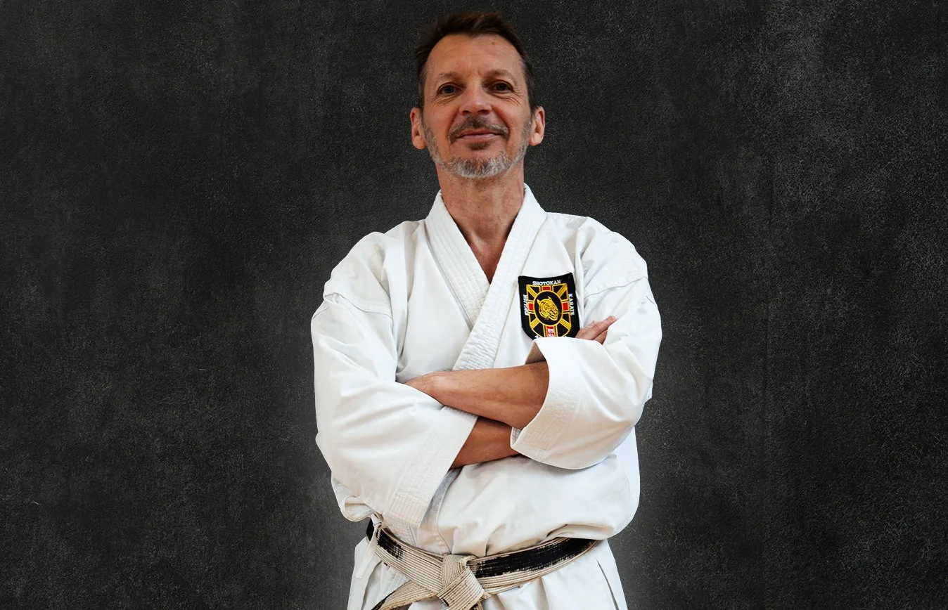 Sensei Warwick Gray, wearing a white martial arts gi with a black belt, standing against a black background