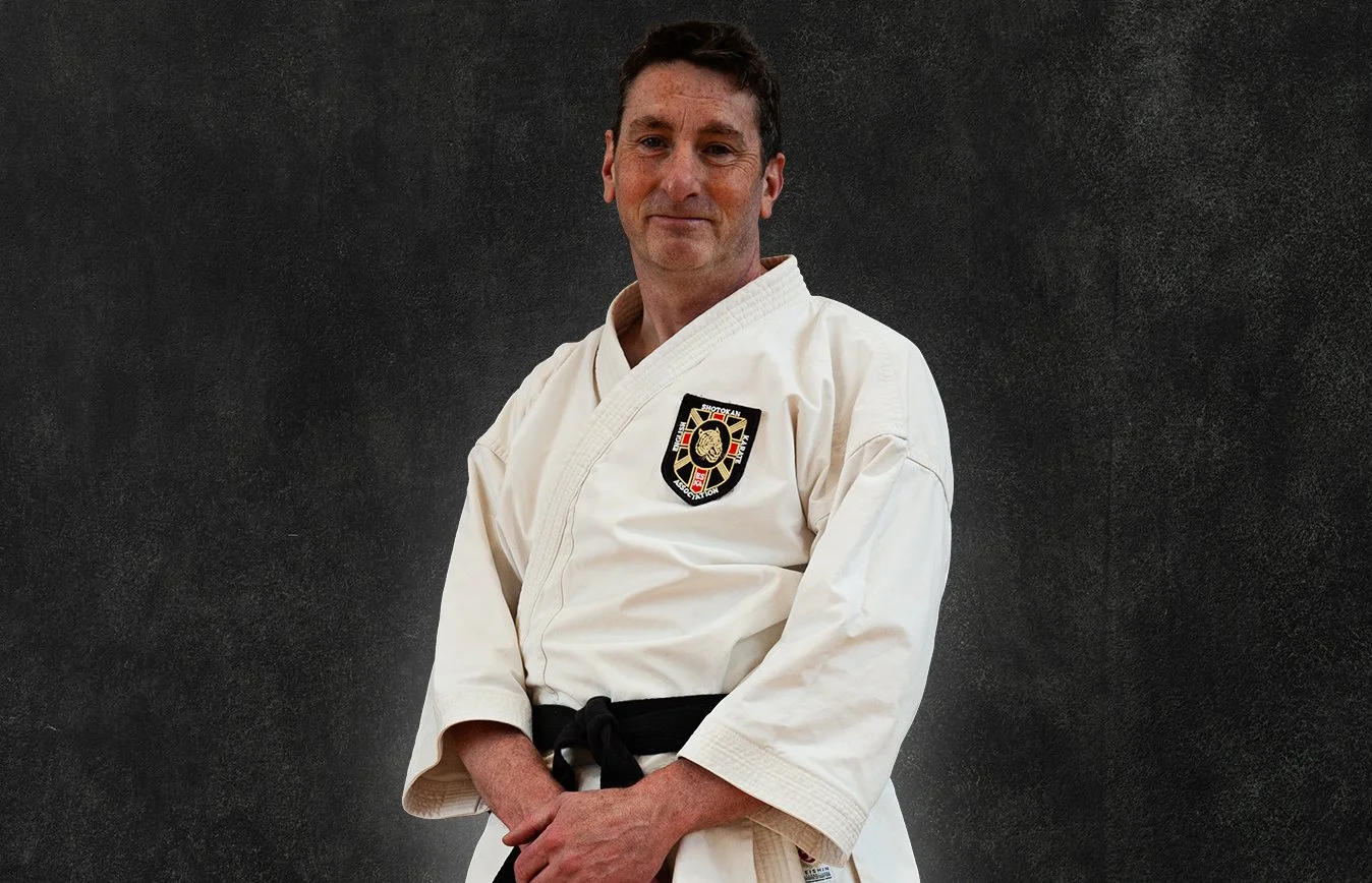 Andrew Leedham, wearing a white martial arts gi with a black belt, standing against a black background
