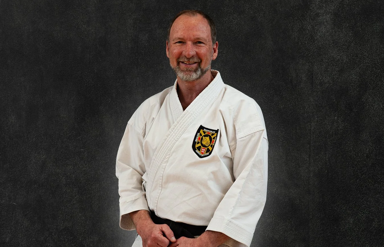 Greg Wood, wearing a white martial arts gi with a black belt, standing against a black background