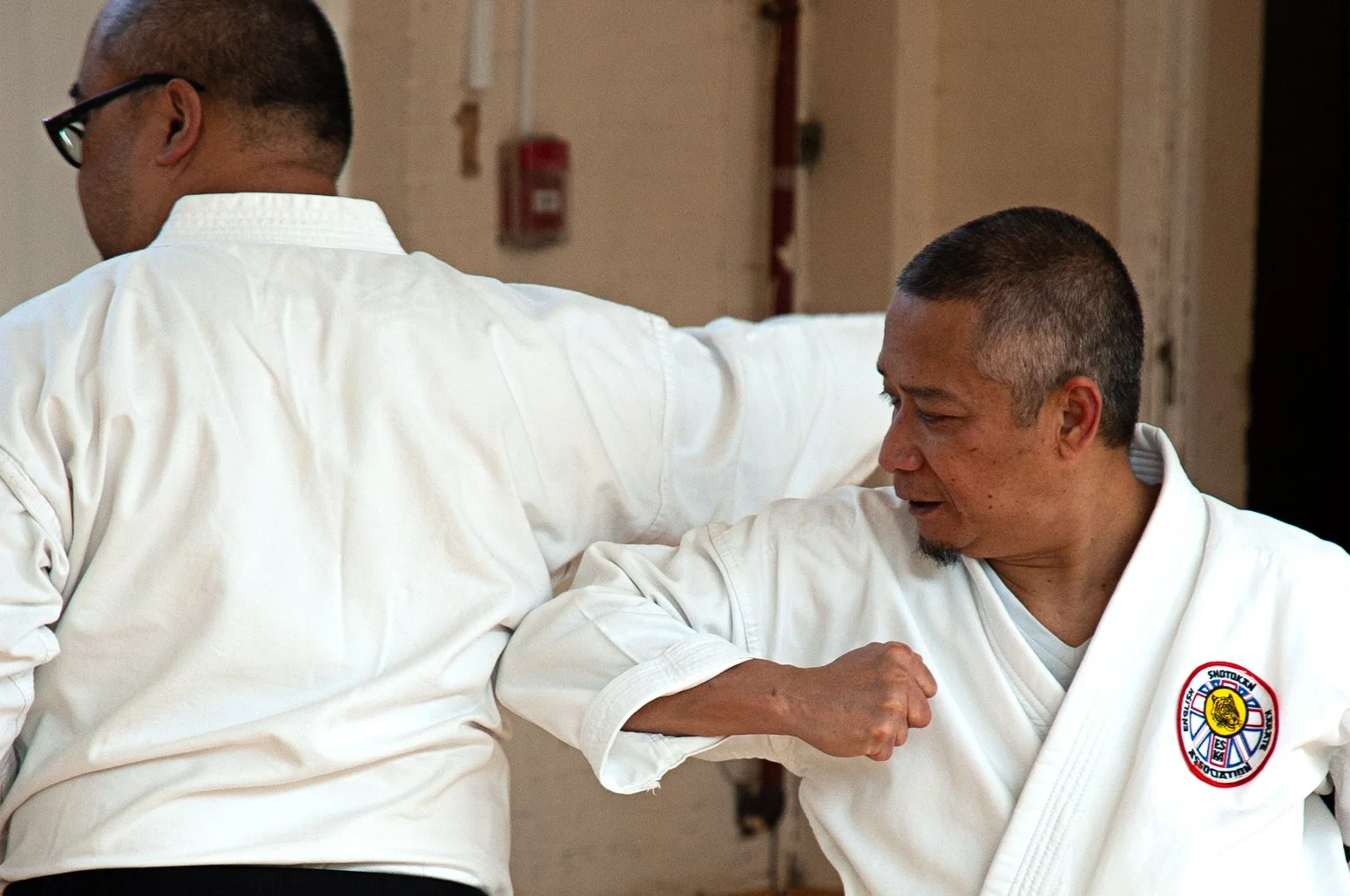 Two men in white martial arts uniforms, with one man striking the second man in the ribs with an elbow technique