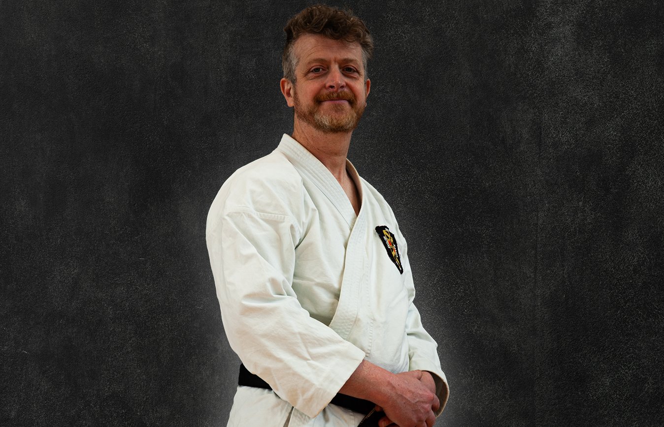Tom Whalley, wearing a white martial arts gi with a black belt, standing against a black background