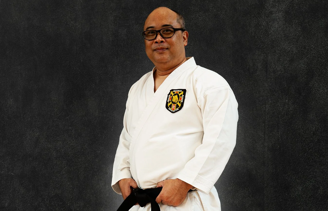 Jenson Basa, wearing a white martial arts gi with a black belt, standing against a black background