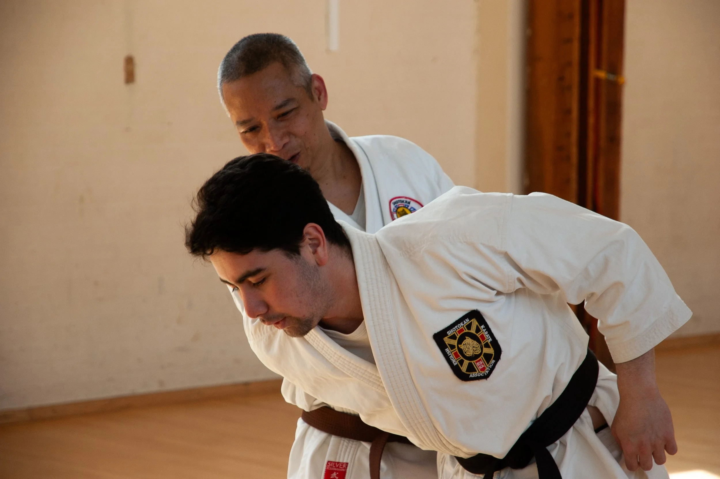 Two martial artists practicing a karate technique on a wooden floor in a training room.