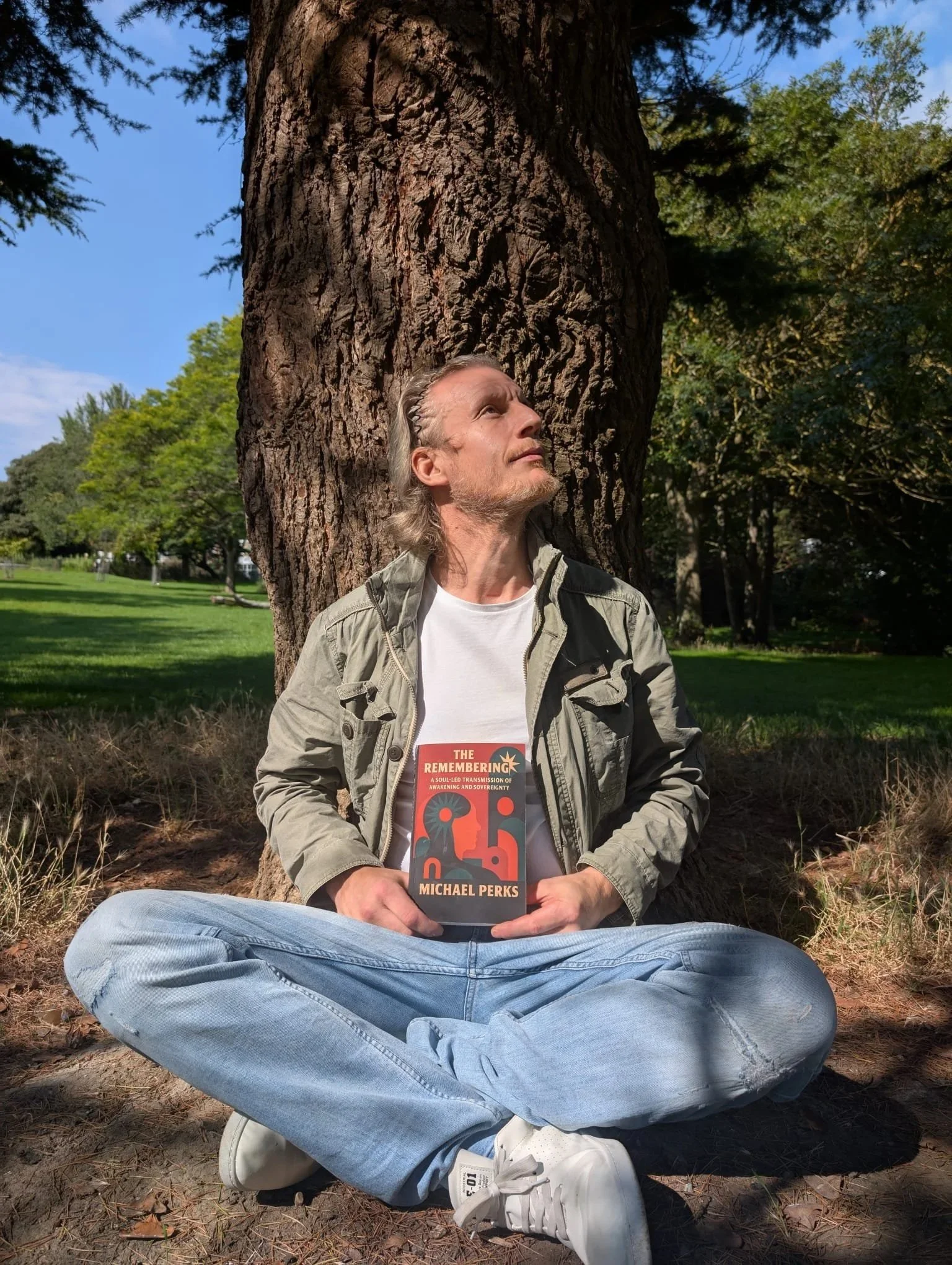 Michael Perks sitting outdoors in nature holding his book The Remembering, representing spiritual awakening, healing, and soul-led transformation.