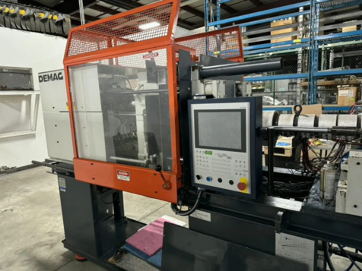 Demag injection molding machine with orange safety guard and control panel, in a warehouse setting with metal shelving and boxes in the background.