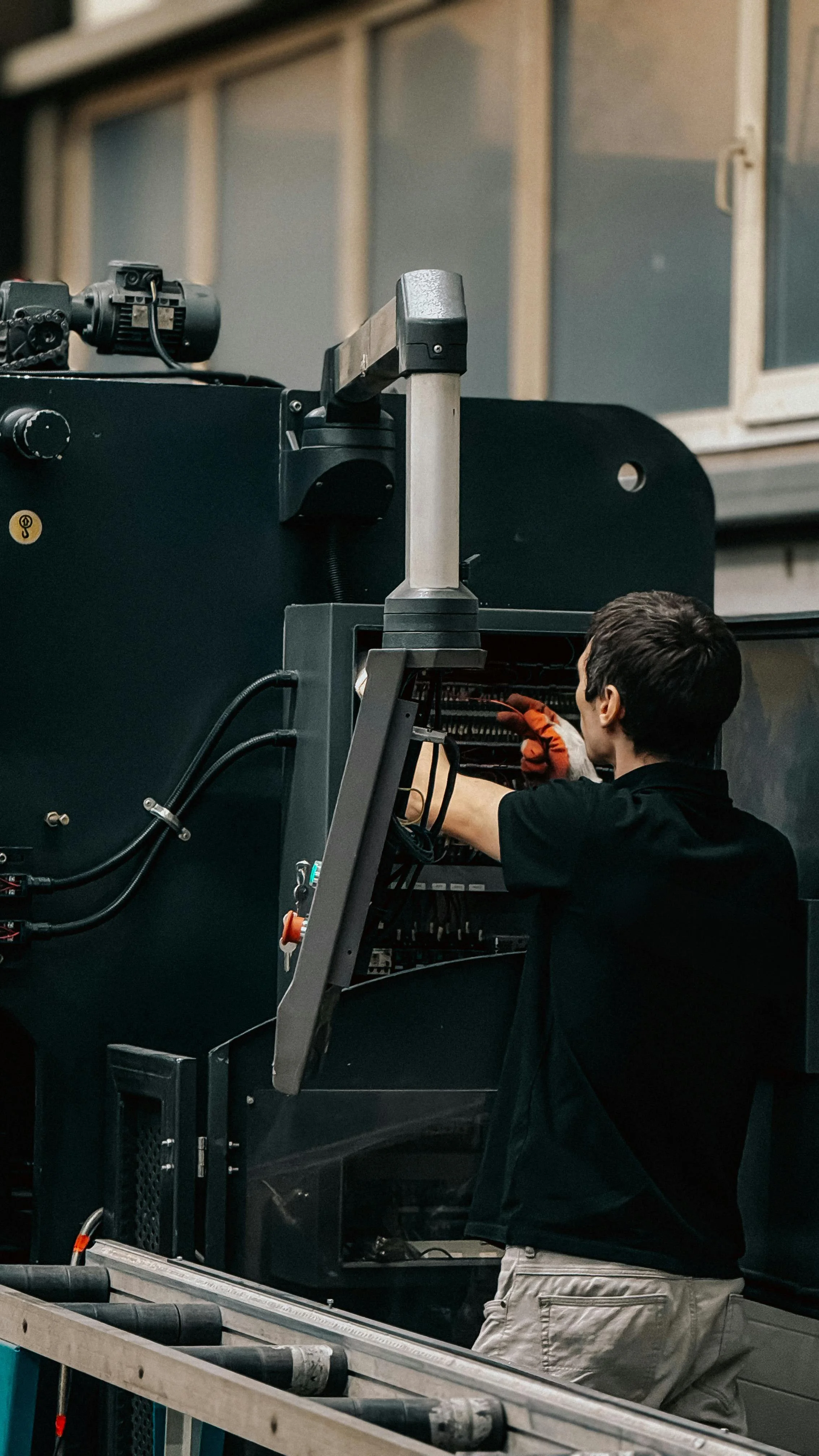 A person wearing a black shirt and beige pants working on a large piece of electronic equipment with wires and components inside. The person is standing in front of the equipment, using tools, in an indoor industrial setting.