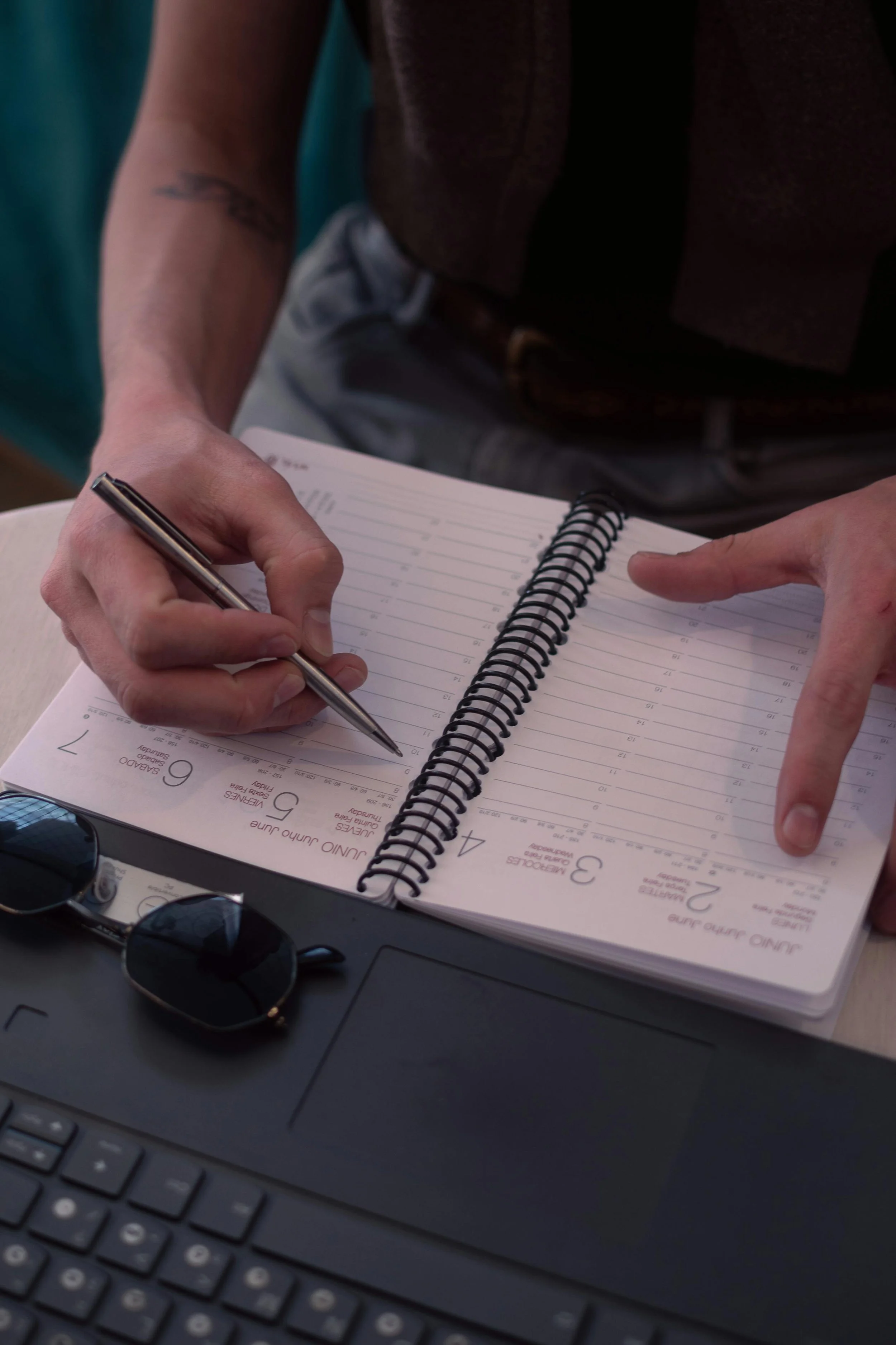 A person holding a pen and writing in a spiral notebook with a calendar page, with sunglasses resting on a keyboard and a laptop visible nearby.