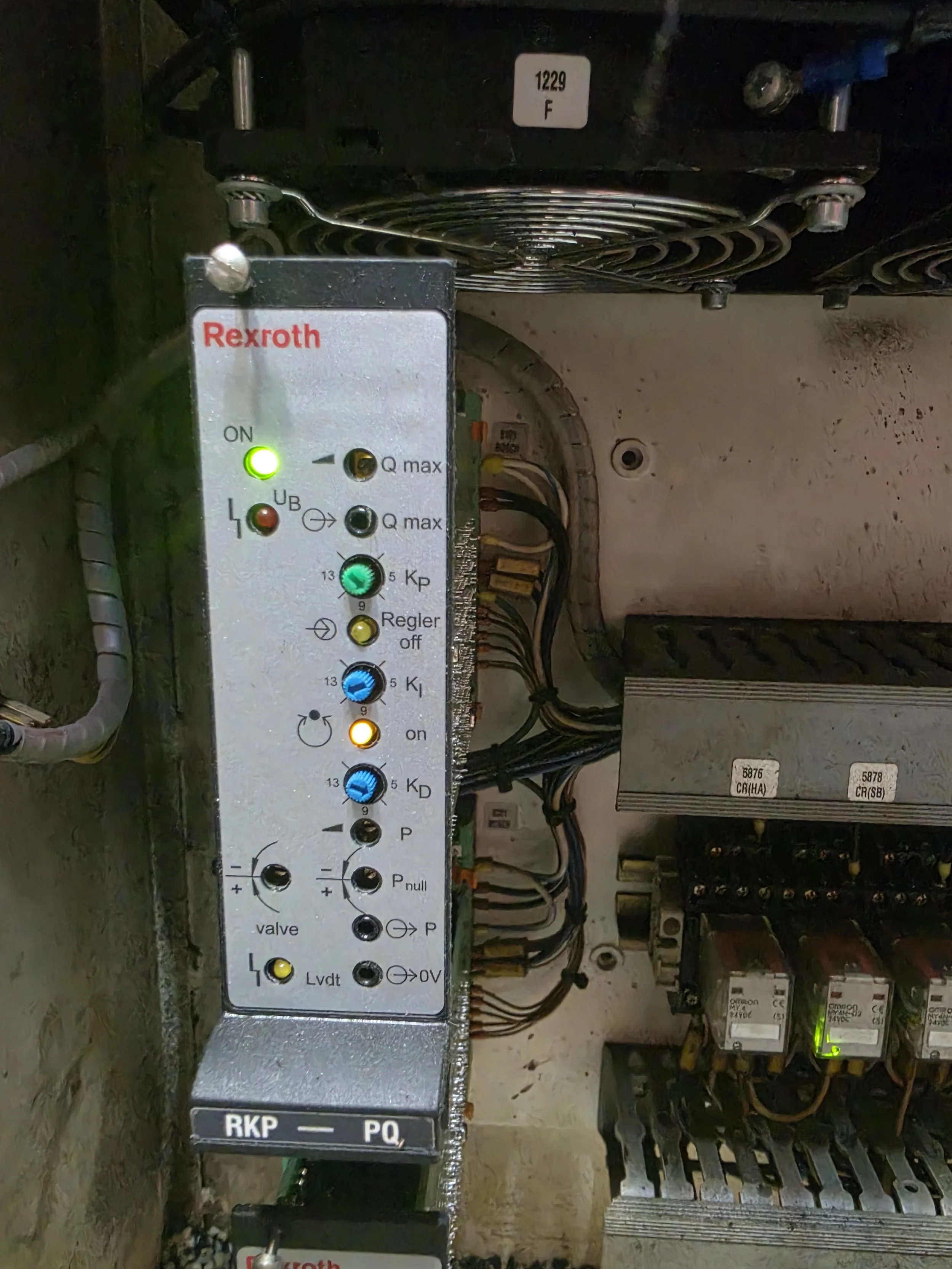 Electrical control panel with various switches, wires, and components. It includes a fluorescent green indicator light, labels, and a brand name 'Rexroth' on a metal plate.