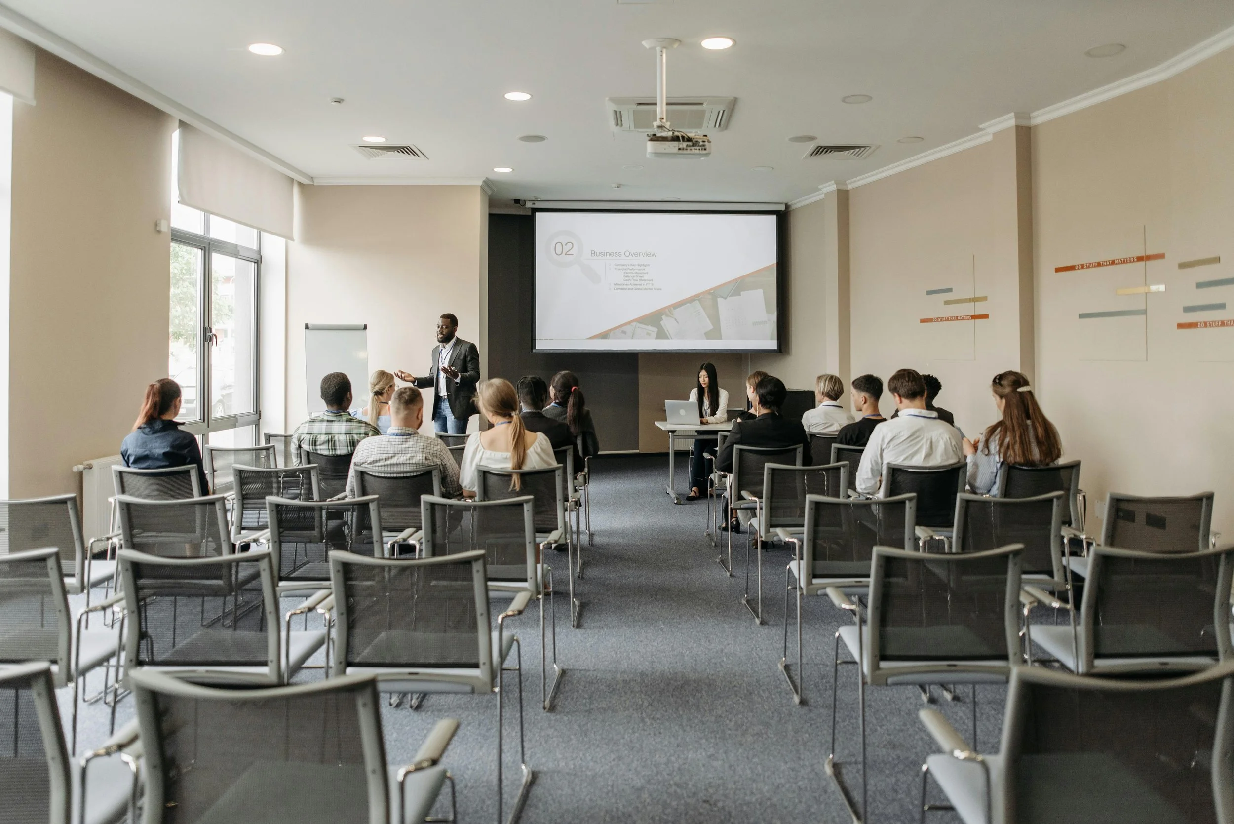 A man giving a presentation in a conference room with a projected slide titled 'Business Overview.' Attendees are seated, facing the presenter, during a professional seminar.
