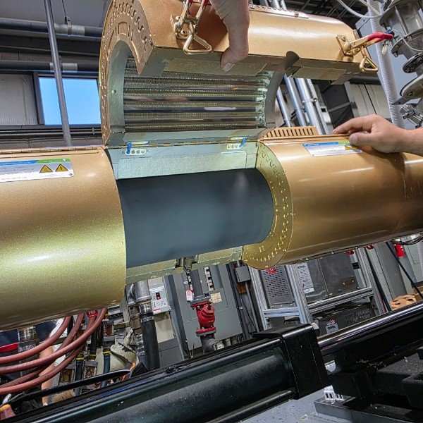 Close-up of a large, cylindrical, gold-colored scientific or industrial apparatus with an opening revealing a dark gray interior, possibly part of a telescope or particle accelerator, with hands adjusting or inspecting it in a high-tech lab or facility.