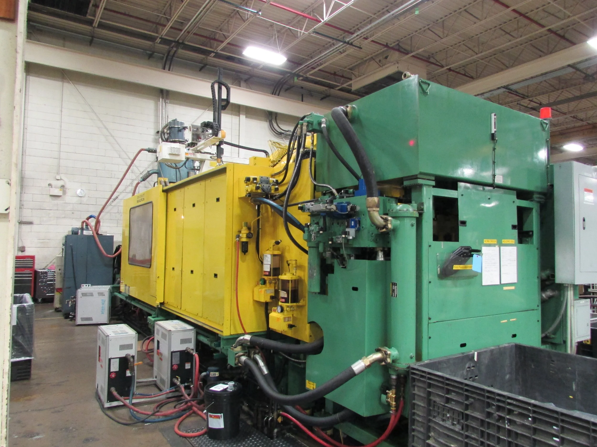 Industrial machine with yellow and green parts, various pipes and cables connected, situated in a warehouse or factory setting.