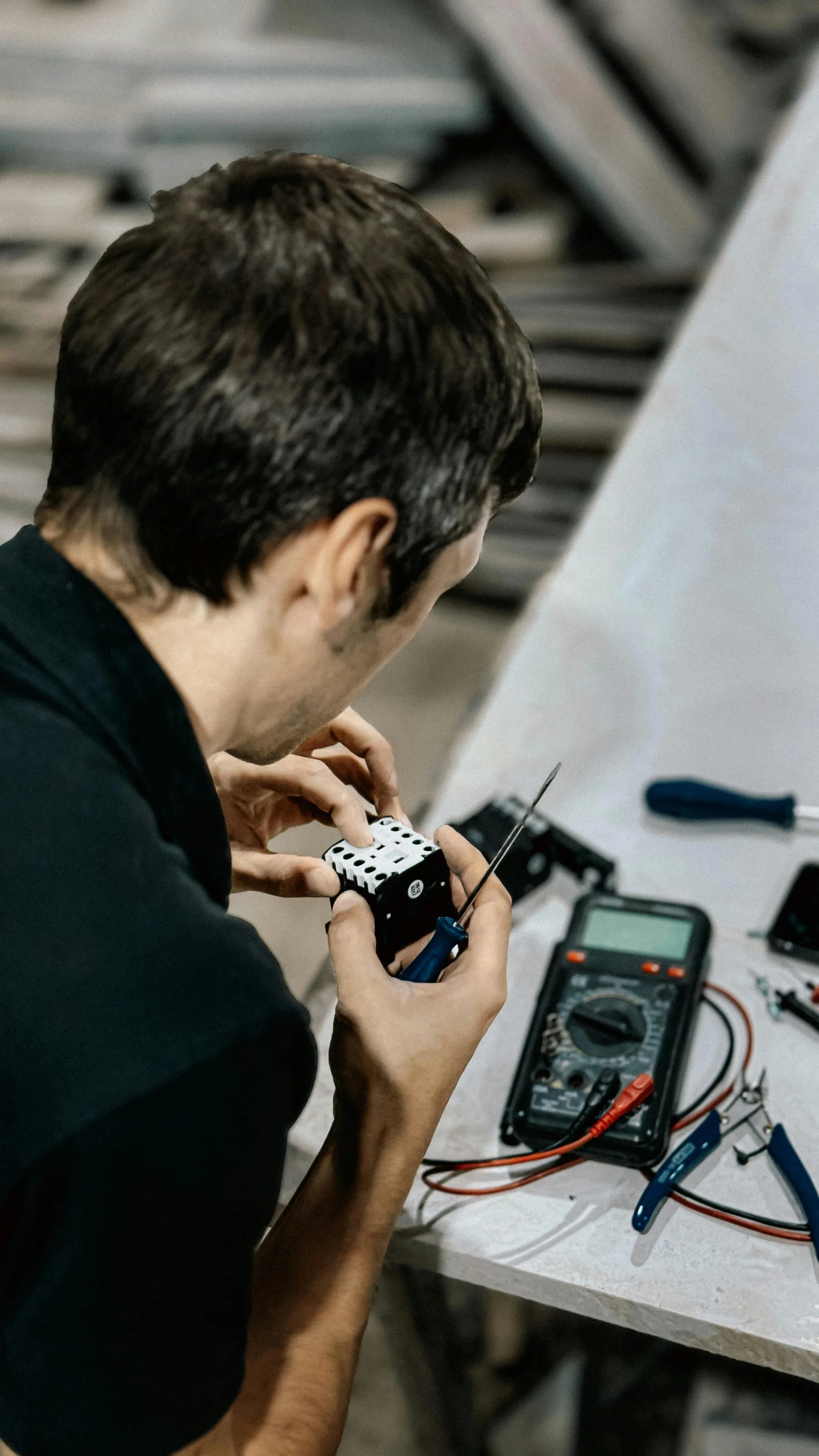 Person working on electronic components with tools, digital multimeter, and circuit boards on a workbench.