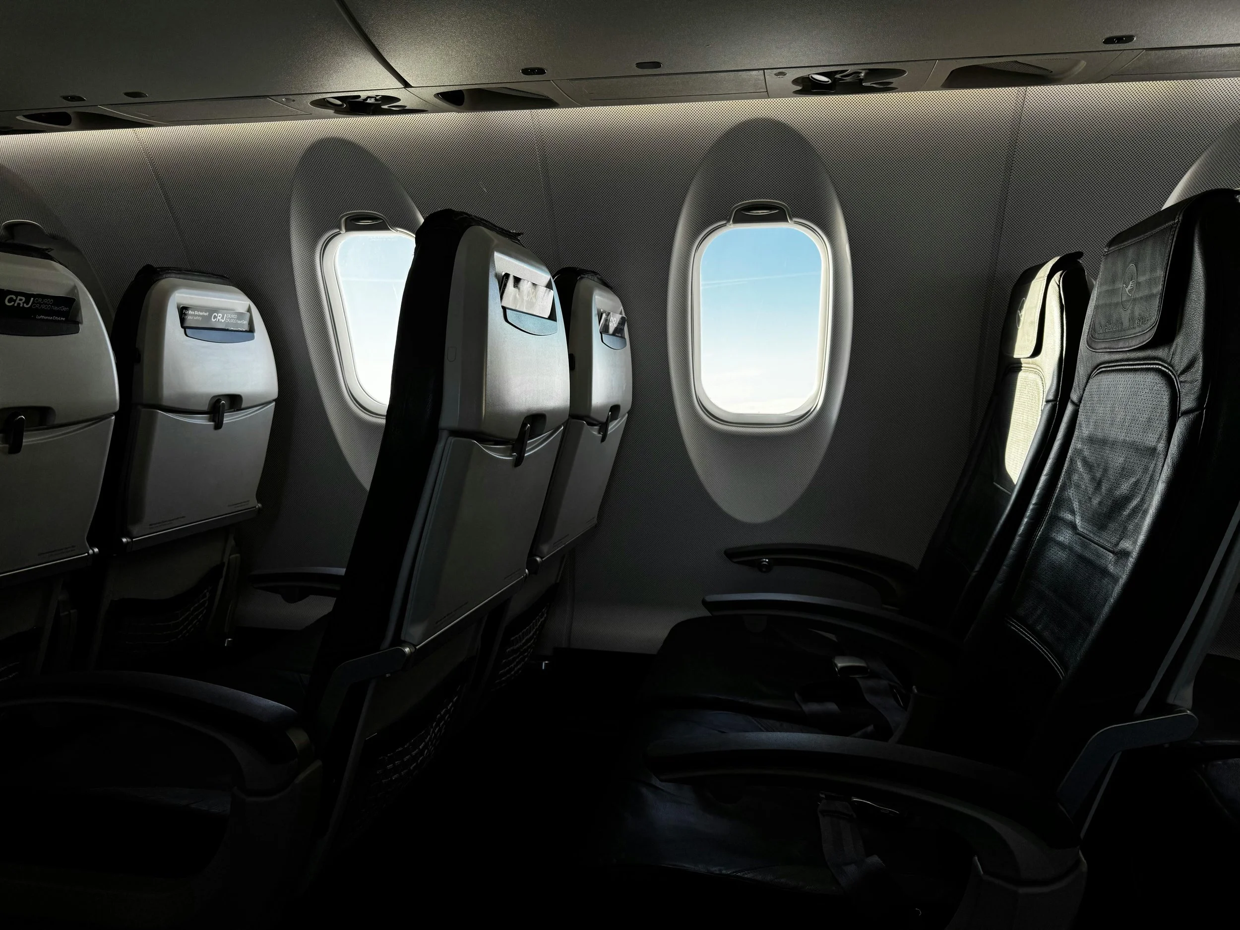 Inside an airplane cabin showing several vacant seats with baggage pockets on the back, two small airplane windows revealing blue sky outside, and a perforated ceiling with built-in overhead controls.