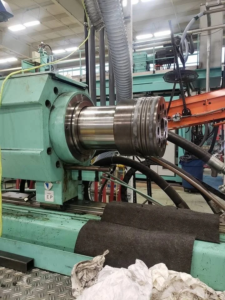 Close-up of a large CNC machine spindle with metal components, surrounded by hoses, wiring, and machinery in a manufacturing setting.