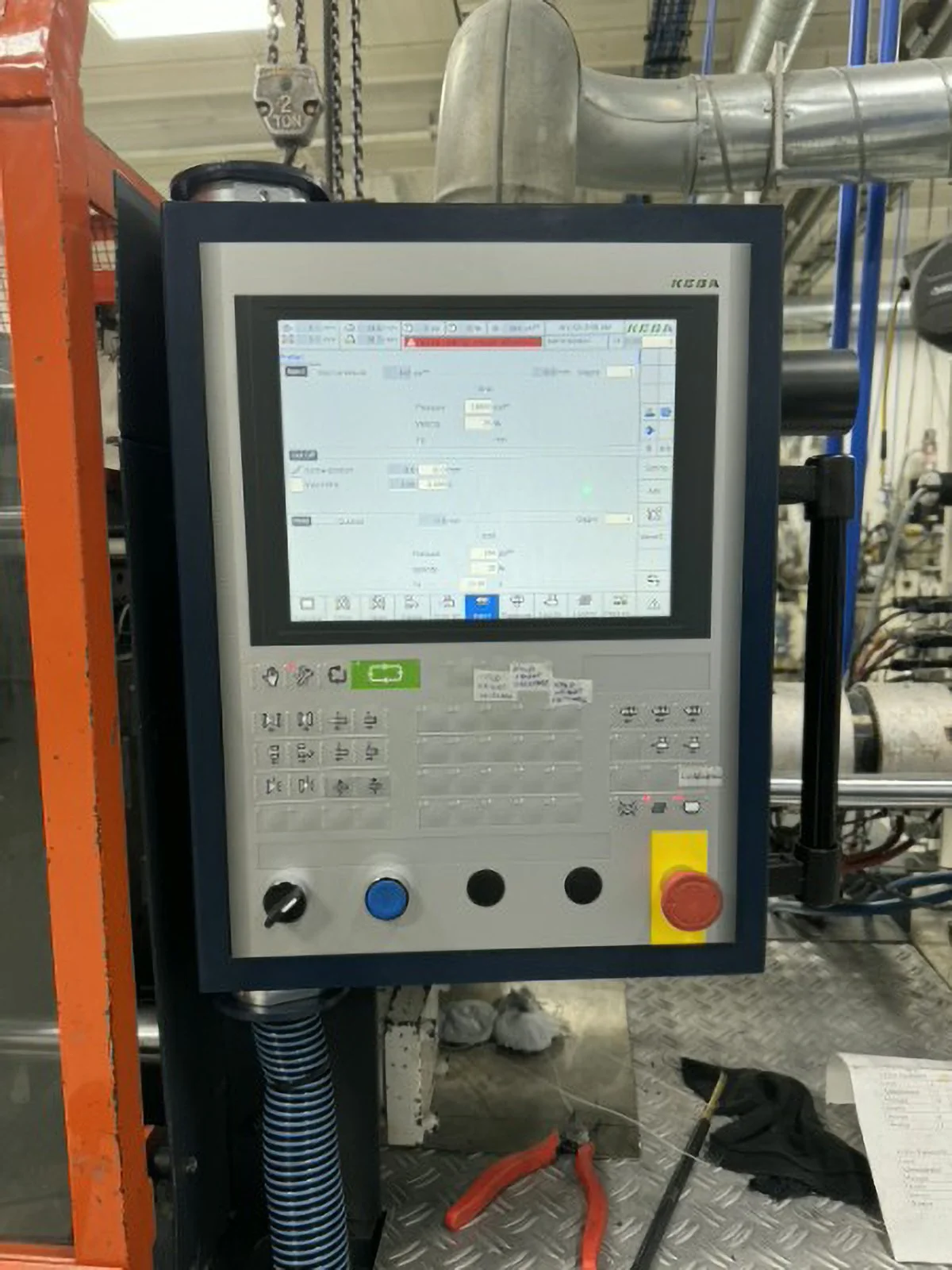 Industrial control panel with screen displaying settings, surrounded by pipes, wiring, and tools in an industrial environment.