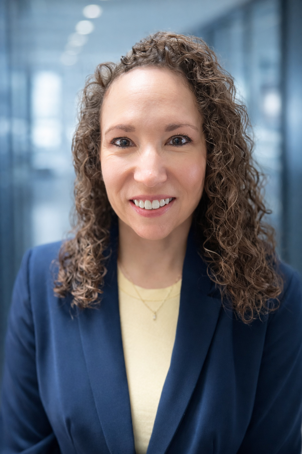 A woman with curly brown hair smiling, wearing a navy blazer and yellow top, standing in a modern office corridor with glass walls.